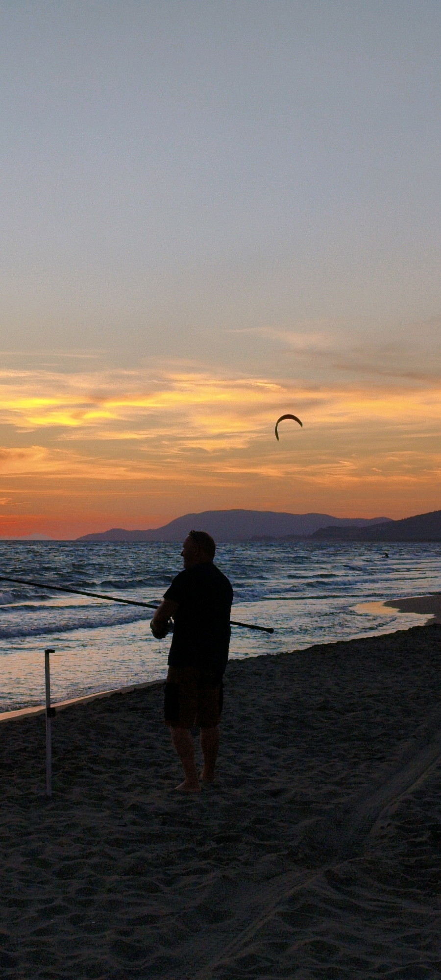 A silhouette of a man is visible from the back, standing on a sandy beach and holding a fishing rod. The sun is setting, casting a warm orange and yellow glow across the sky, with streaks of clouds. In the distance, a kite is flying. The sea is a deep blue with gentle waves. The man is wearing a dark shirt and shorts, and appears to be fishing. The scene captures a serene moment at sunset on a beach, possibly in the Maremma area of Grosseto, Italy, known for its coastal landscapes.