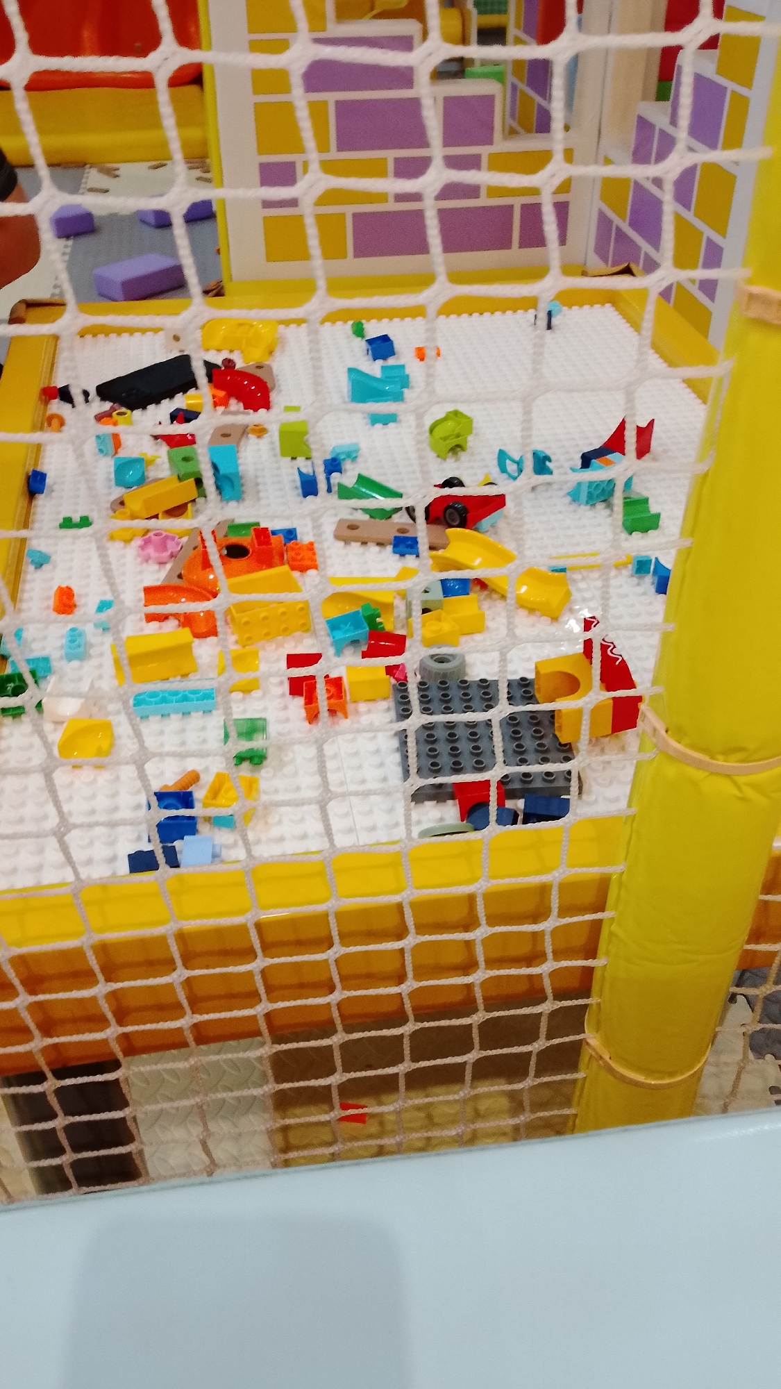 The image shows a close-up view of a children's play area, likely a ball pit or soft play structure. The foreground is dominated by a white netting that partially obscures the scene. Inside the netting, a white mat surface is covered with a variety of colorful plastic building blocks, resembling LEGOs. These blocks are scattered and assembled into various small structures. Dominant colors include yellow, orange, red, blue, green, and purple. In the background, there are larger, brightly colored structures with geometric patterns, primarily in yellow and purple. A thick yellow cylindrical pole is visible on the right side of the image, wrapped with a light brown band. The overall impression is a playful and colorful environment designed for young children. The location is specified as a children's play area in South Tangerang, Indonesia. There are no people visible in the image.