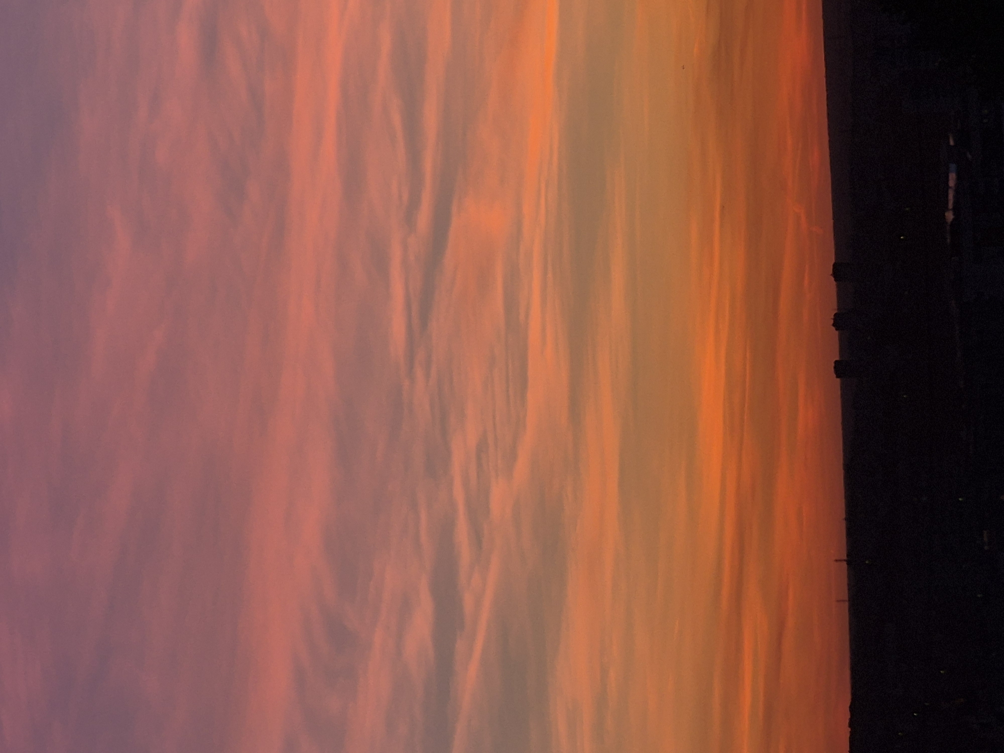 The image depicts a vibrant sunset over an urban area in an unknown city. The sky is filled with clouds, exhibiting a gradient of colors from pink and purple on the left to orange and yellow on the right. On the right side of the image, a dark silhouette of buildings and structures is visible, indicating the city skyline. The angle is slightly off, as the picture is rotated counter-clockwise.