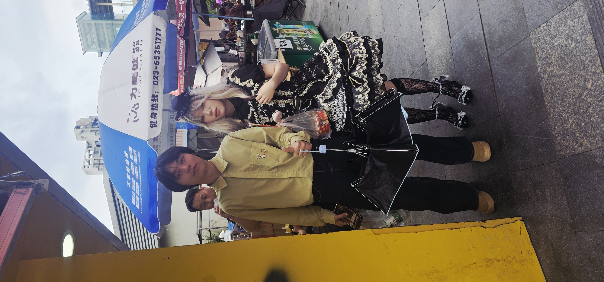 The image shows two people posing on a sidewalk near an outdoor market in the city of Chongqing High-tech Zone, China. A fair-skinned woman with blonde hair wearing a black and gold lolita dress with lace stockings and black heeled shoes reclines on the shoulder of a man. The man holds a closed, black umbrella. He wears a light yellow collared shirt, black pants, and tan shoes. Under the blue and white awning of an outdoor market, another man stands behind the pair. The market extends along the sidewalk with various vendors displaying wares. The sidewalk is paved with grey tiles, and a yellow curb is in the foreground. Text in Chinese and English indicates businesses advertising fitness and educational services. The buildings of Chongqing High-tech Zone are visible in the background.