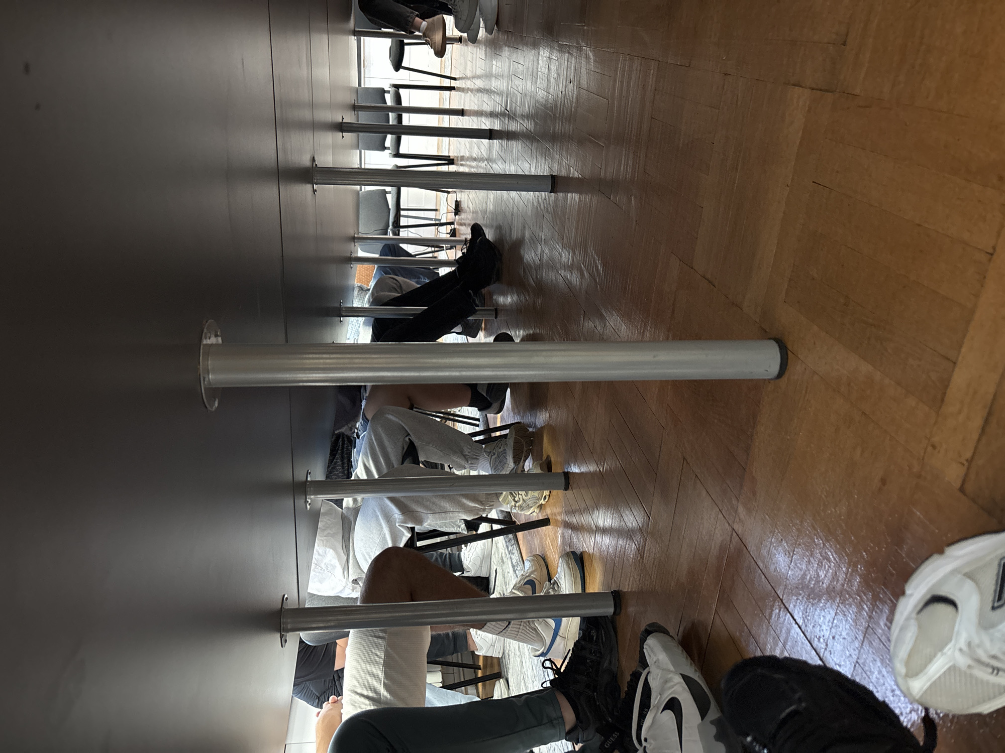Under a long, dark table with metal legs, multiple people sit on chairs. Their legs and feet are visible beneath the table. The floor is made of wooden planks. This likely takes place in a public space in Zlatograd, Bulgaria, given the prompt's request.