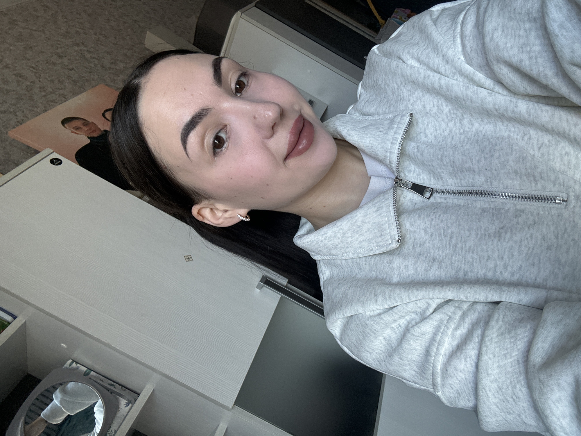 A woman with dark hair and fair skin, wearing a light gray zip-up sweatshirt, is posing for a close-up, slightly tilted selfie. The background includes a light-colored wooden surface with drawers and shelves, and an out-of-focus picture of a man on a wall. The photo may have been taken indoors, possibly in the woman's home or apartment in Марковский, Russia, although this cannot be verified from the image alone. The woman has a neutral to slightly smiling expression, and her makeup is visible.