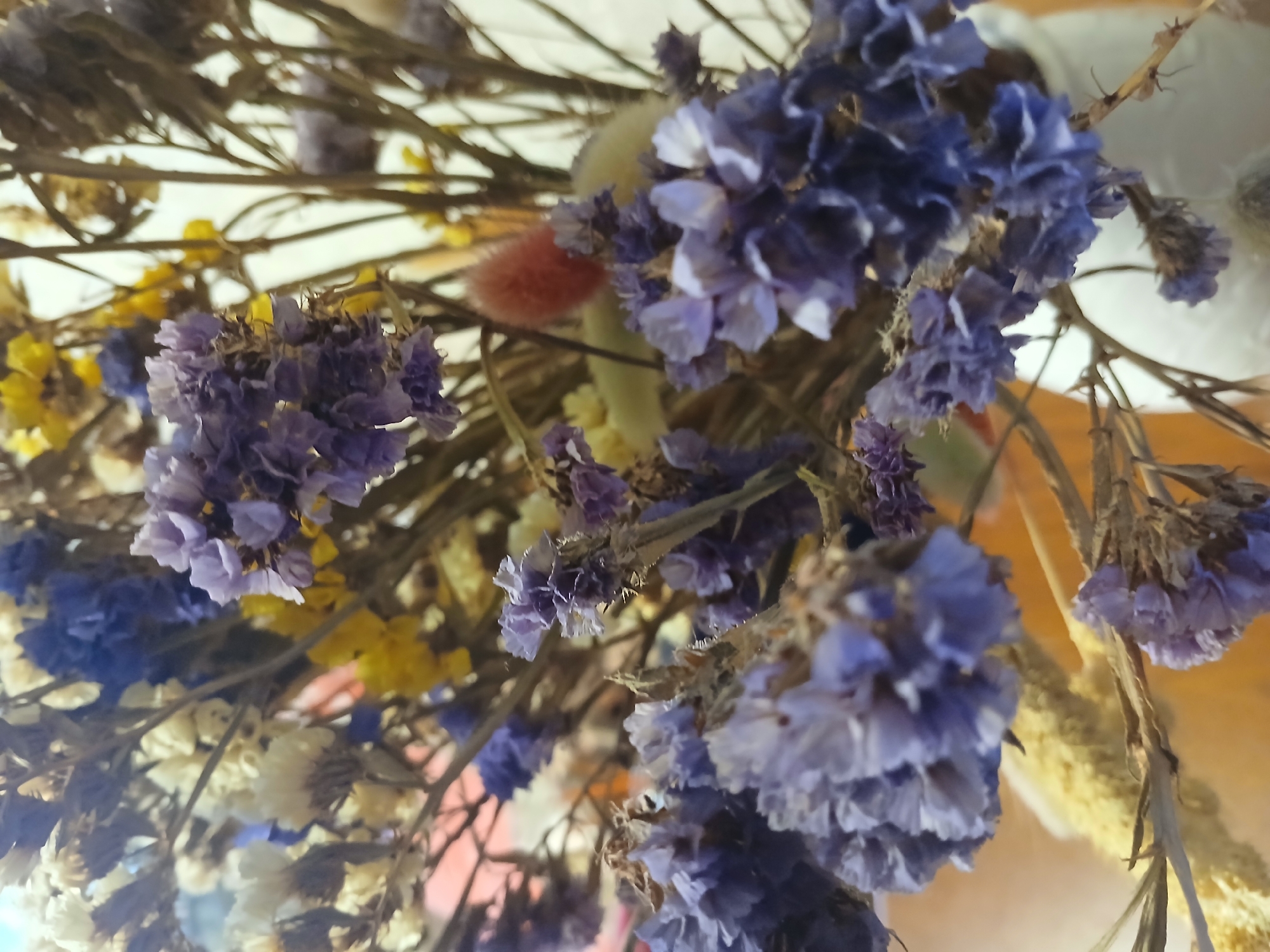 The image shows a close-up view of a bouquet of dried flowers, including varieties with small purple, white, and yellow blossoms. There are also some fuzzy, decorative elements in orange and green. The background is blurred, but there are hints of a light-colored surface or wall. There is no discernible information to identify the location as Kamianske, Ukraine or any other city. The image does not depict any people, actions, or interactions.