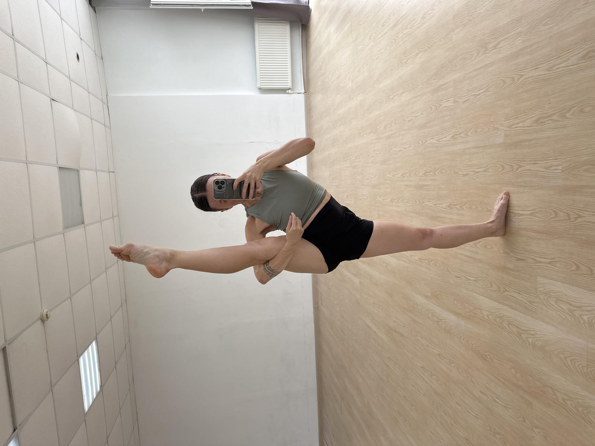 A woman is performing a vertical split in a brightly lit room with a light-colored wooden floor. She is wearing a grey tank top and black shorts. She is holding a smartphone in her right hand, appearing to take a selfie. The room has white walls, a dropped ceiling with fluorescent lighting panels, and a white radiator against the back wall. There is a large mirror on the left side of the room, reflecting the scene. There are no visible clues to indicate the location is in Votkinsk, Russia.