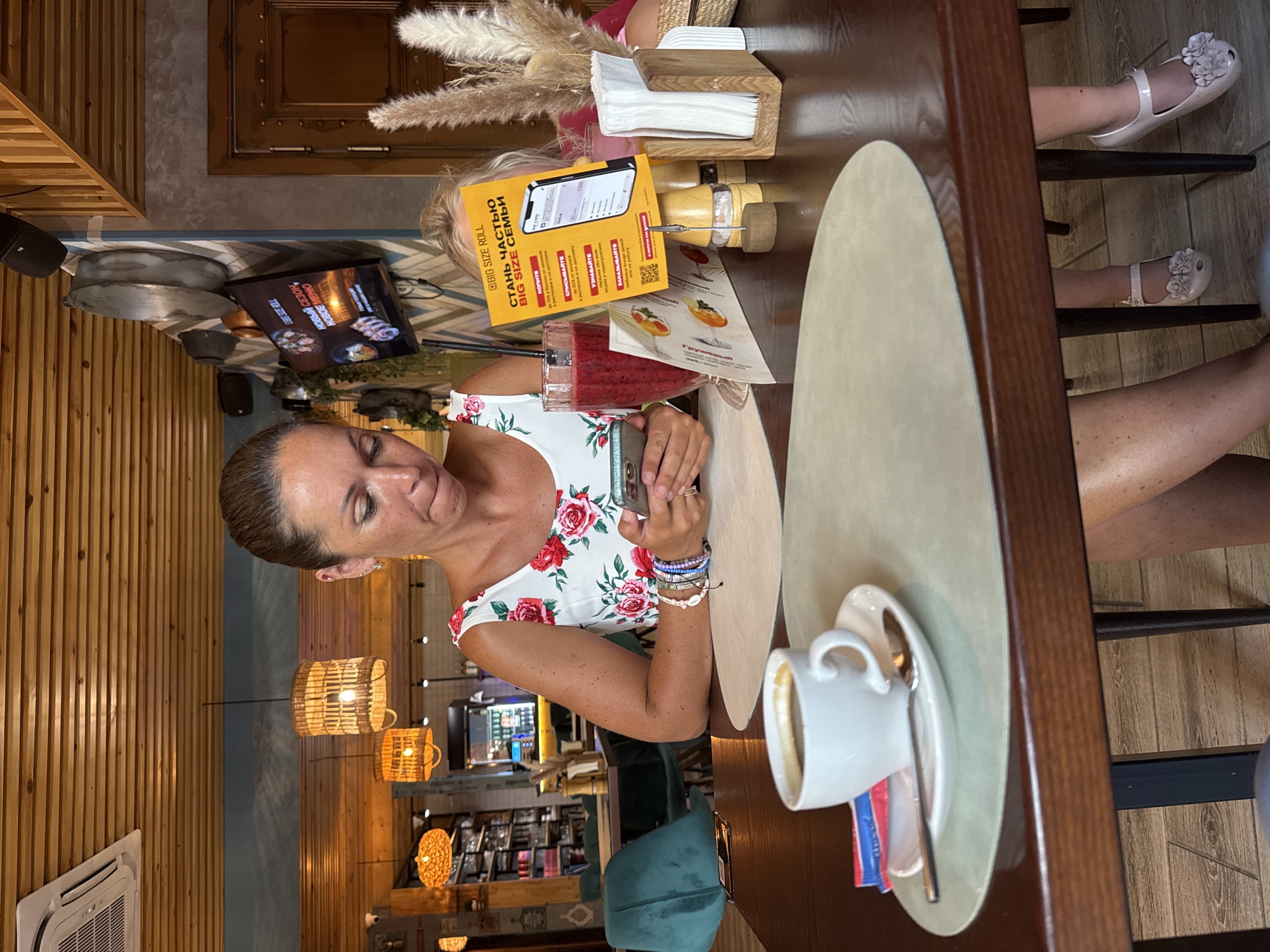 The image depicts an indoor scene within a restaurant in Sochi, Russia. A woman is seated at a table, looking at her mobile phone. She is wearing a floral print dress and several bracelets. In front of her is a cup of coffee, a spoon, and a sachet of sugar. To her right, a glass of red smoothie and a menu stand are visible. Another person, partially visible, is seated across from her. The restaurant features wooden ceiling beams, rattan light fixtures, and a wall decorated with a chevron pattern.