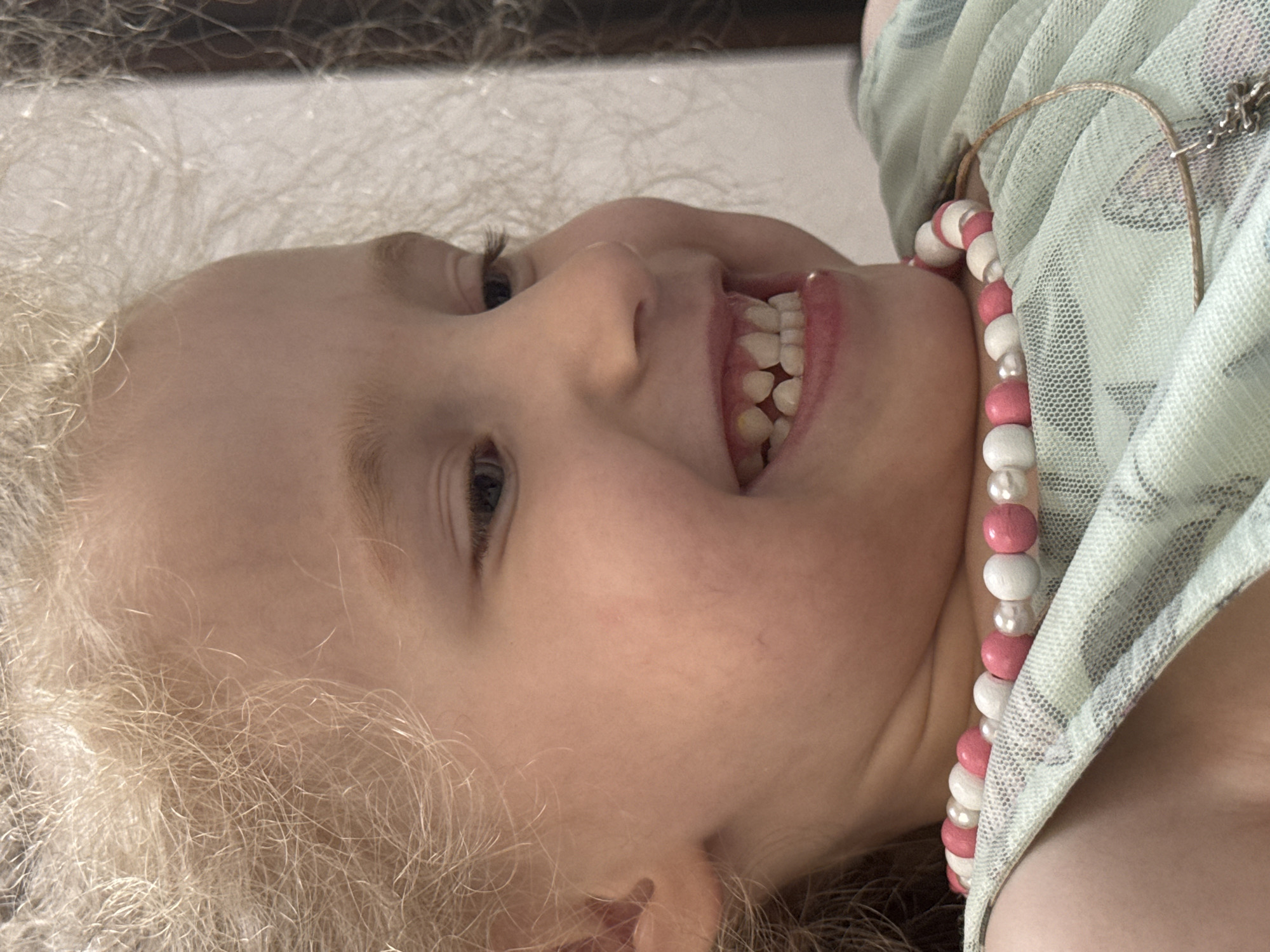 A close-up, waist-up, profile shot shows a young girl with blonde, curly hair and a light complexion, wearing a light green, sheer, patterned top and a pink and white beaded necklace. She is smiling, revealing her teeth, and looking to her right. The background is out of focus, but appears to be an indoor setting with a light-colored wall and possibly some dark vertical elements. The image is well-lit, with the girl's face as the primary subject. The location is not discernible from the image.
