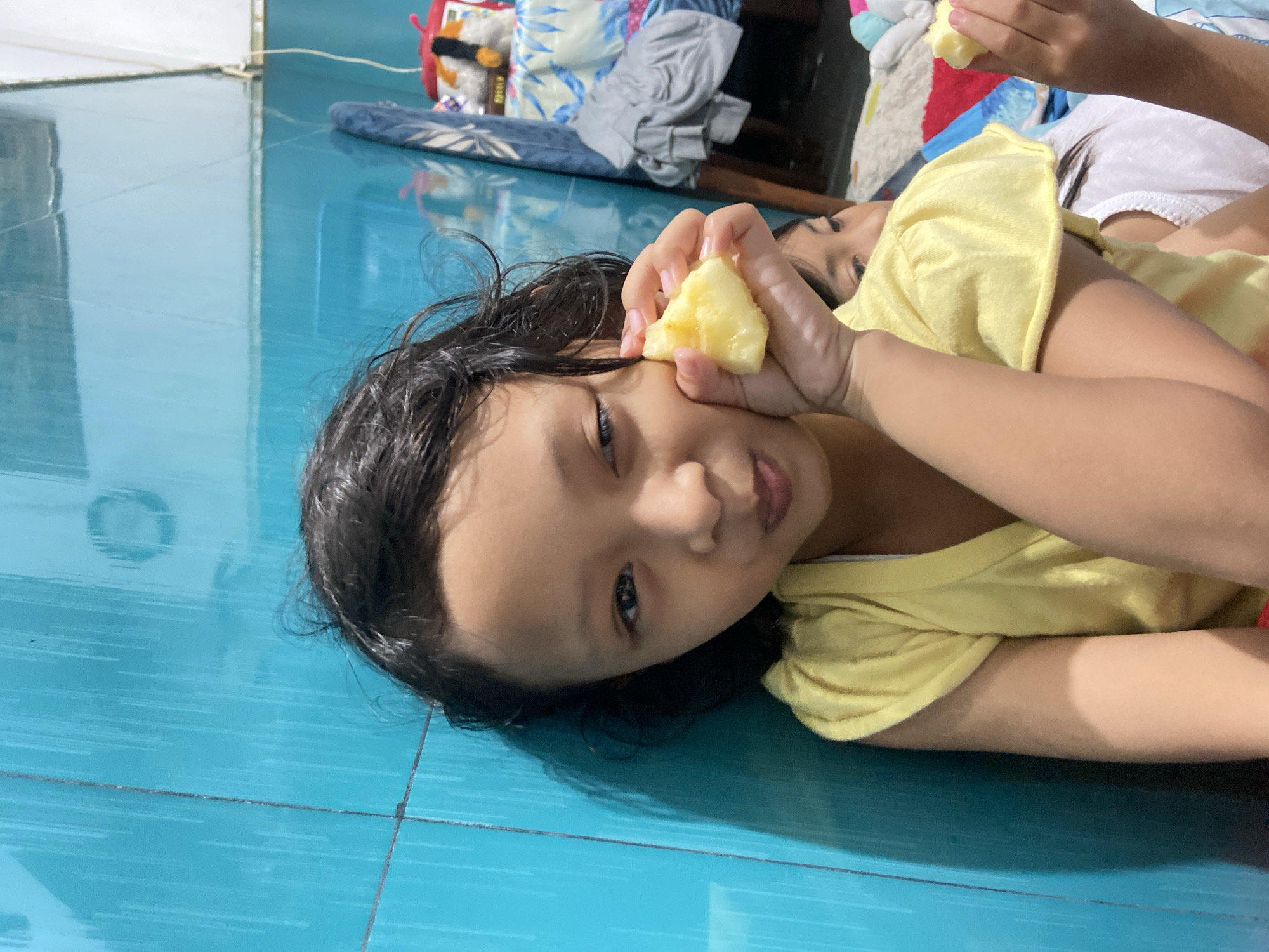 A young Asian child with dark hair is lying on a glossy blue tiled floor. She is wearing a yellow shirt and is holding a piece of yellow fruit near her face, possibly eating it. Another child, partially visible, is lying beside her. The background includes a patterned blanket and assorted items. The setting appears to be indoors, possibly in a home. The location is Gelam, Indonesia.