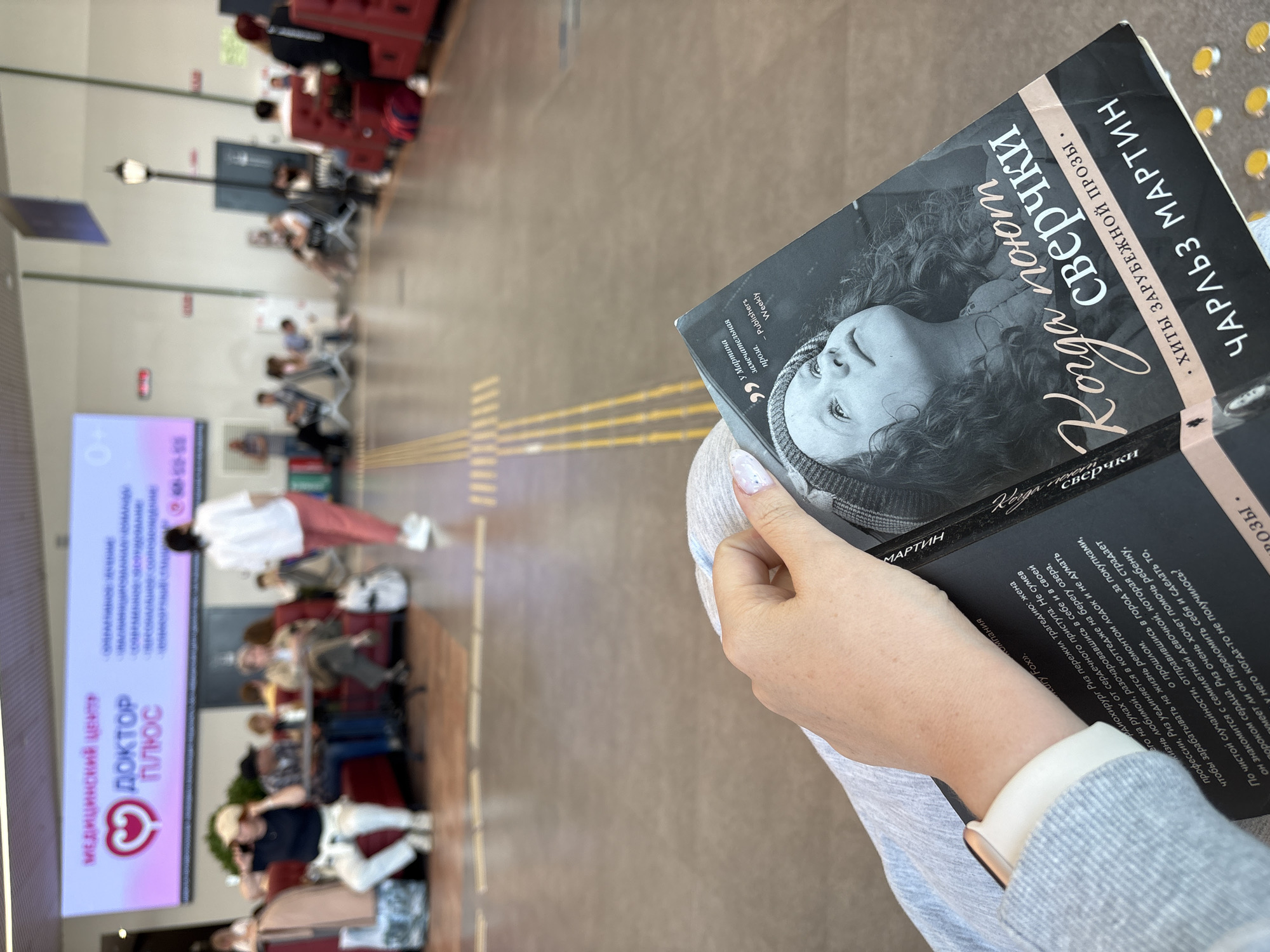 Here is a concise description of the image:
The image shows a person sitting in what appears to be a waiting area, possibly an airport or medical center, holding a book titled "Когда поют сверчки" (When the Crickets Sing) by Charles Martin. The book is a Russian translation. In the background, there are people seated in chairs, a digital display with information, and other elements suggesting a public waiting area. The book is in focus in the foreground, while the background is slightly blurred. Based on the OCR of the book, it is possible this location is near or in Новое Мартьяново, Russia.