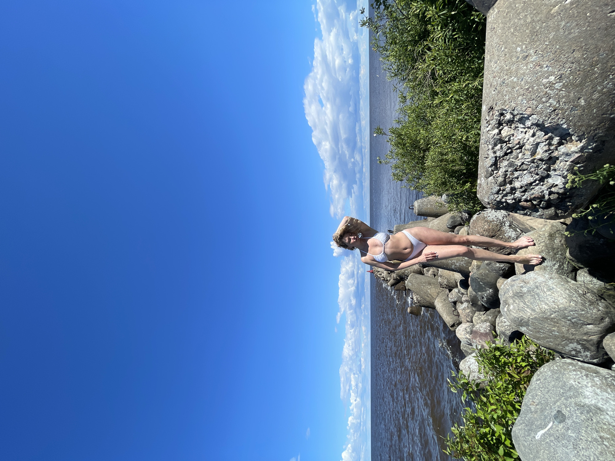 In a full shot taken outdoors, a woman stands on a rocky embankment, a body of water to her left, and a clear blue sky with sparse clouds above. She is positioned on a large grey boulder, wearing a white bikini, with her left hand touching her hair. The woman's pose is relaxed, with her weight shifted to her right leg. The embankment is made up of large, rough grey rocks and concrete structures. Green foliage is visible on the right side of the image, growing among the rocks. The water appears dark and slightly rippled. The overall impression is a sunny day in a coastal or lakeside environment, possibly a breakwater or pier. The location is the city of Belozersk, Russia, a historic city on the shores of Lake Beloye.