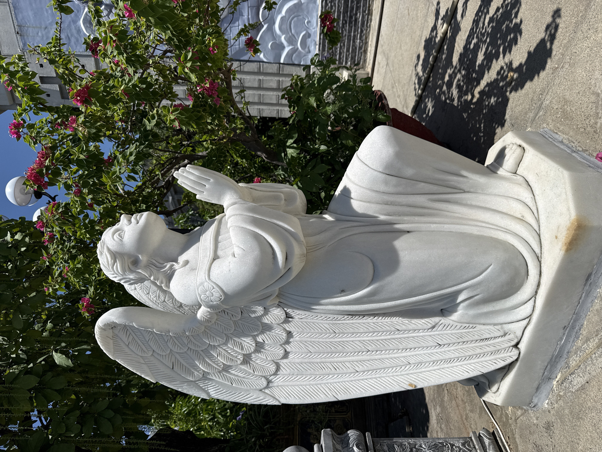 The image depicts a white marble statue of an angel kneeling with its hands clasped in prayer, looking upwards. The statue is positioned outdoors, with a lush green tree adorned with pink flowers and a blue sky in the background. To the right of the statue, a light gray wall with a decorative cloud motif is visible. The statue is situated on a paved ground surface, casting shadows from the surrounding foliage. The scene appears to be in a garden or courtyard, possibly within a religious or spiritual site in Nha Trang Ward, Vietnam, given the tropical vegetation and architectural style.
