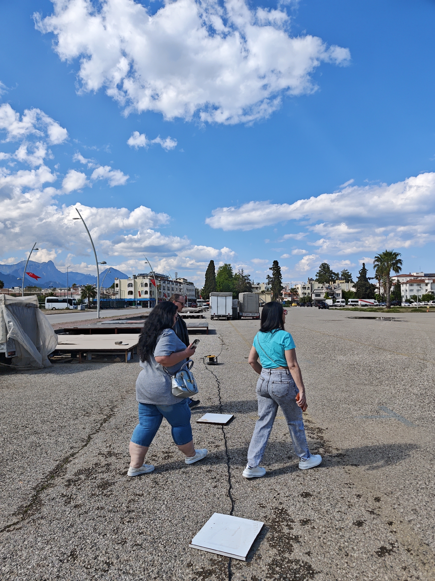 The image depicts two women walking across a parking lot in Kemer, Turkey. One woman is wearing a grey top and blue jeans. The other is wearing a blue shirt and grey jeans. The parking lot is mostly empty with some equipment including what looks like large flat tiles and some covered equipment. In the background are buildings, mountains, and a Turkish flag. The sky is blue with white clouds.