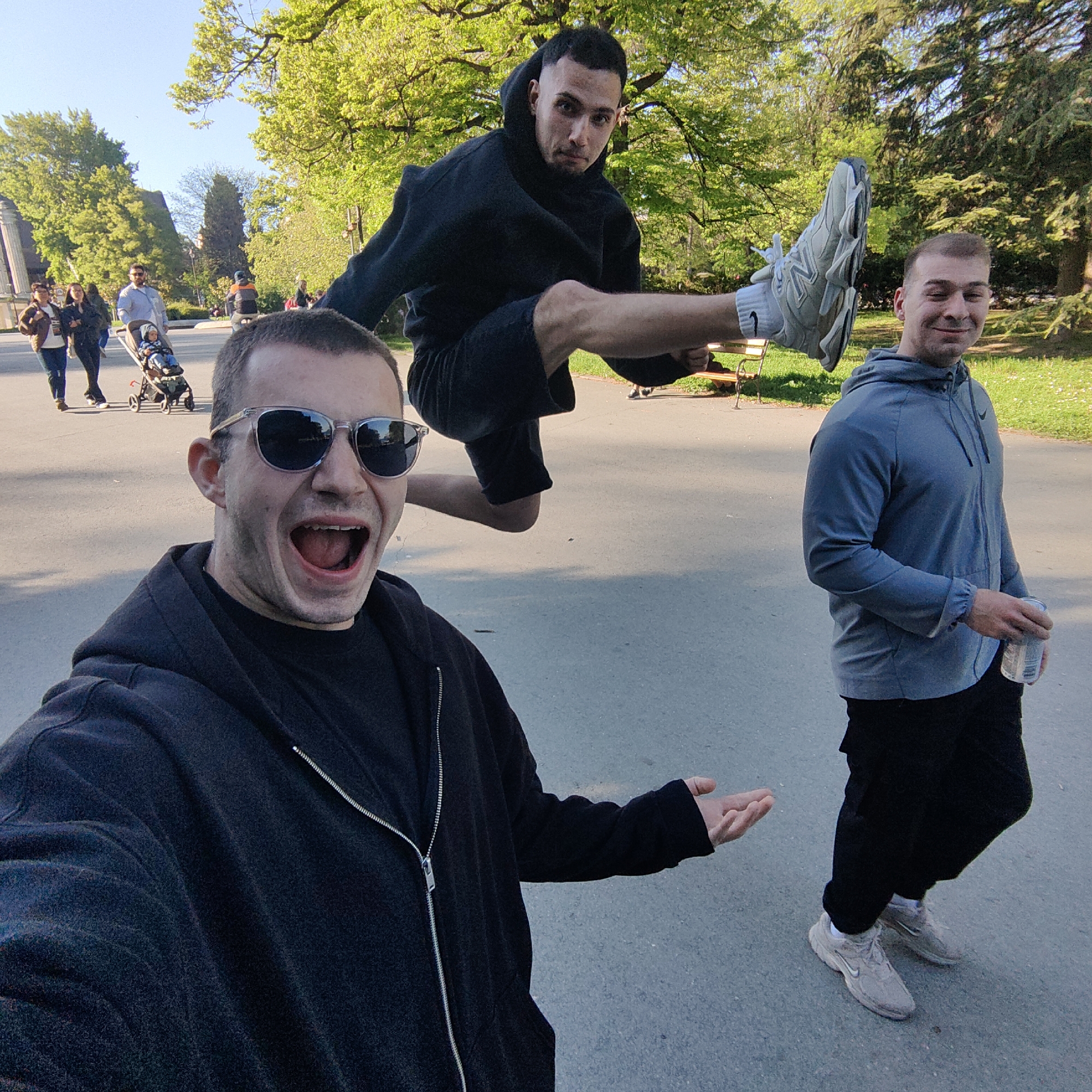 The image appears to be a selfie taken in a park in Varna, Bulgaria. Three men are present. The man taking the selfie is in the foreground wearing sunglasses and a black zip-up hoodie. He has his mouth open wide and is gesturing with his right hand. Behind him, a second man is suspended mid-air, appearing to jump or kick, wearing a black hoodie and shorts. To the right of the jumper, a third man stands on the ground, holding what appears to be a canned drink, wearing a grey hoodie and dark pants.
In the background, other people are walking on a paved path, including a couple with a stroller. There is a stone building visible in the distance, along with trees, grass, and a park bench. The setting suggests a public park during daylight hours.