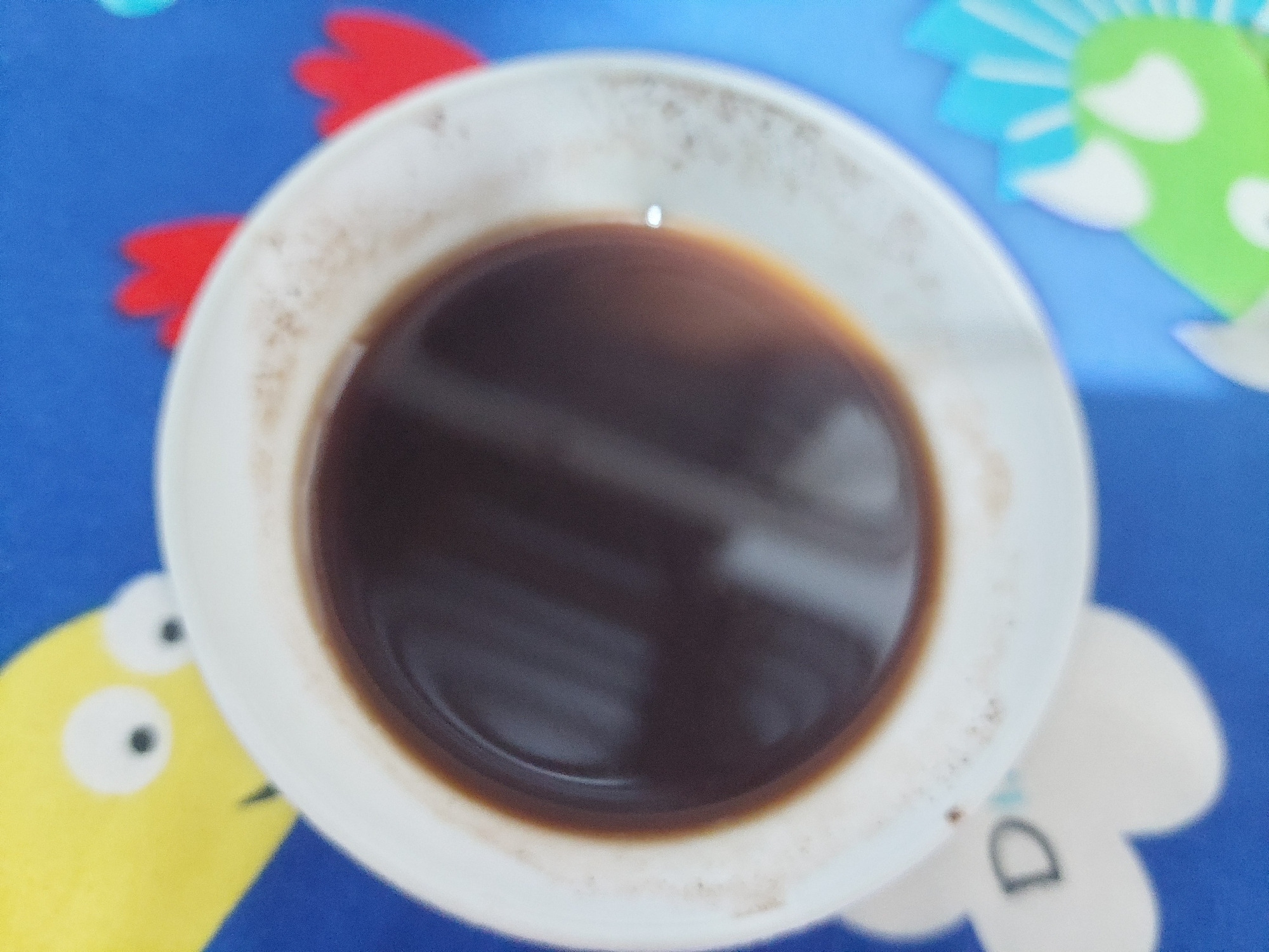 A close-up, blurry, top-down shot shows a white ceramic mug filled with dark liquid, likely coffee. The mug has some brown stains around the rim. The mug is placed on a blue surface that appears to be a placemat or tablecloth with colorful cartoonish figures, including a yellow character with two eyes and some red and green cartoonish shapes. A small, bright reflection is visible on the surface of the liquid. The scene suggests a casual setting, possibly a home or cafe, in Jelutung, Indonesia. There is no visible text in the image. The lighting is diffused, and the overall impression is one of a simple, everyday moment.