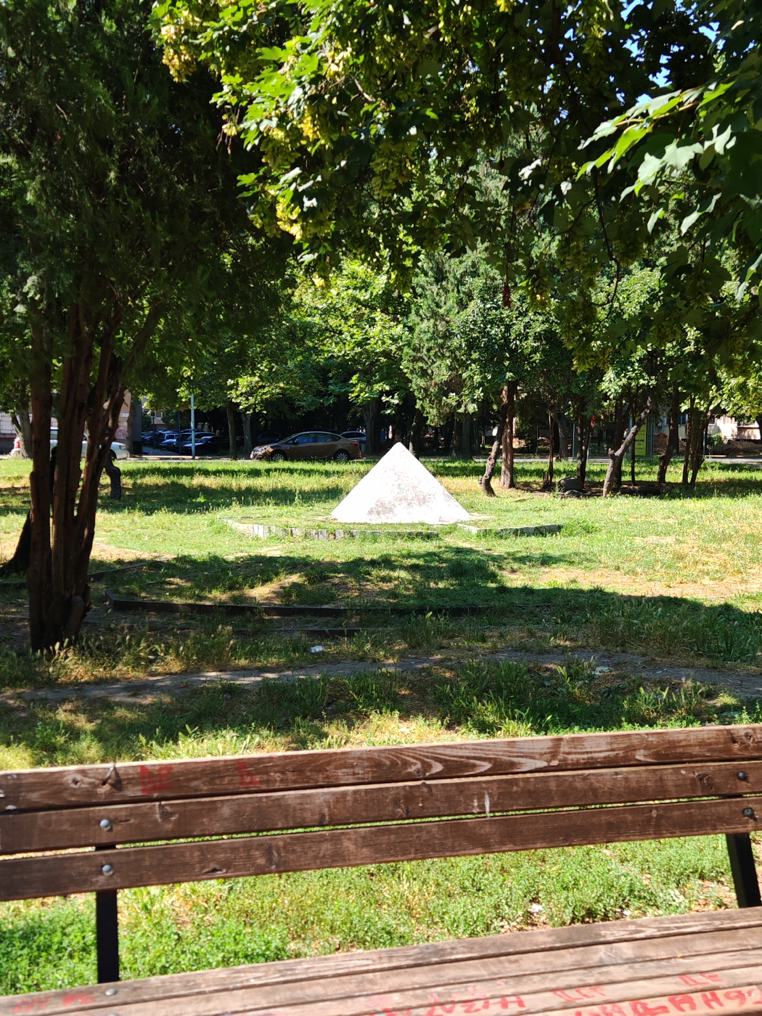 The image depicts a serene park scene in Plovdiv, Bulgaria, bathed in the warm glow of midday sunlight. The foreground is dominated by a weathered wooden bench, its surface marked with graffiti, offering a stark contrast to the lush green grass and scattered wildflowers beyond. A paved path, partially visible, separates the bench from a manicured lawn, where a geometric, pyramid-like structure stands as a solitary monument.
The midground is a verdant expanse of grass, shaded by the dense foliage of mature trees that form a natural canopy, dappling the ground with shifting patterns of light and shadow. In the distance, a few cars are parked along a street, hinting at the proximity of urban life. The trees, with their thick trunks and abundant leaves, create a sense of enclosure, drawing the viewer's eye towards the central pyramid. The sky, a clear, bright blue, is partially obscured by the overhead branches, suggesting a pleasant, warm day.