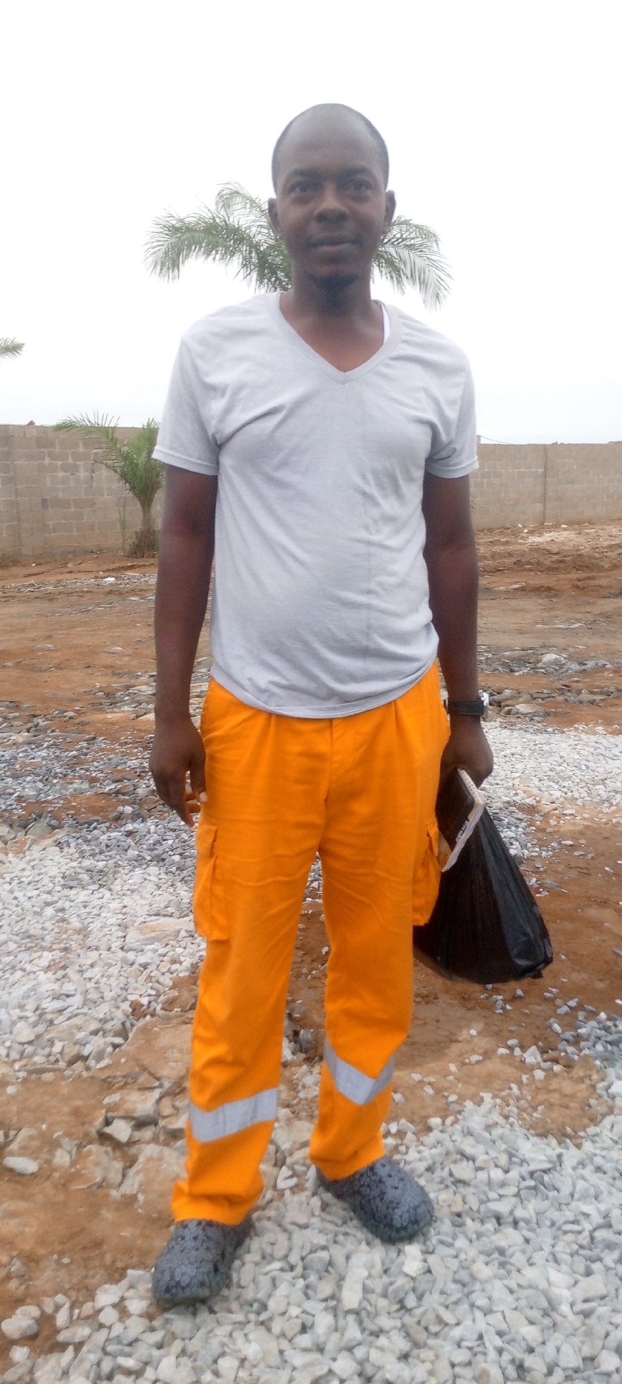 A full-body, slightly low-angle portrait captures a Black man standing outdoors in what appears to be a construction site. The man is wearing a light gray V-neck t-shirt and bright orange cargo pants with reflective silver bands around the lower legs. He has dark skin, a shaved head, and is looking directly at the camera with a neutral expression. He is holding a black plastic bag in his right hand. The background shows a low brick wall, some sparse vegetation, and uneven, dusty ground covered in gravel. The sky is overcast. The scene is likely in Indo, Nigeria, given the context.