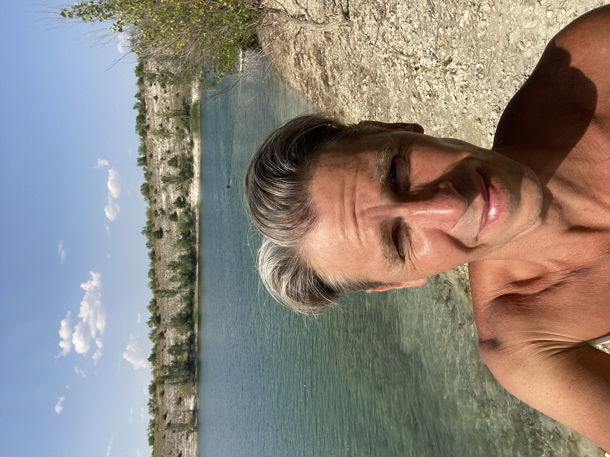 The image is a selfie taken outdoors. A shirtless man with grey hair is in the foreground, smiling at the camera. He appears to be at a beach or lakeside. The background shows a body of clear, turquoise water with a rocky shore. Beyond the water, there is a slope covered in dry vegetation and scattered trees, under a bright blue sky with scattered clouds. The location is identified as Куйбышевское сельское поселение, Ukraine. There is a person swimming in the water in the distance.