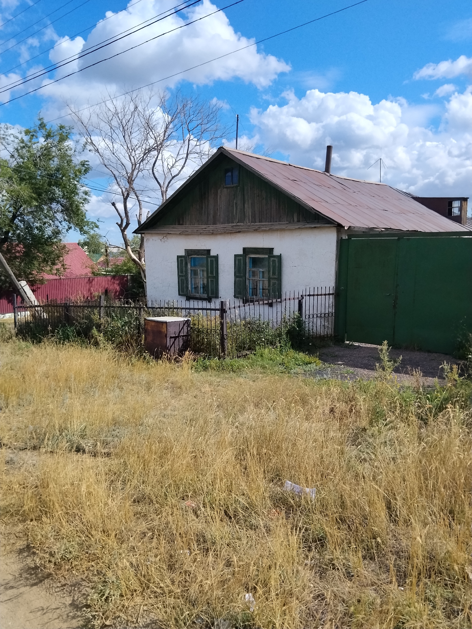 The image shows a simple, white, single-story house with a rusted metal roof and wooden siding. The house has two open windows with green shutters. A green metal gate is attached to the right side of the house, and a low black metal fence encloses a small area in front of the house. The yard in front of the fence is overgrown with dry, yellow grass. A rusty metal barrel is visible inside the fenced area. Behind the house, a red fence and more houses are visible, suggesting a residential area. The sky is blue with scattered white clouds, and power lines are visible in the upper portion of the image. The overall scene depicts a rural or semi-rural residential property. The image is likely from Kokshetau, Kazakhstan, given the context provided.