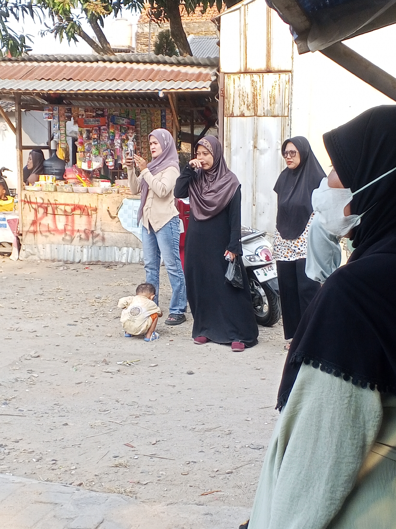 A group of people are standing and crouching near a small roadside stall in Ciruas, Indonesia. In the foreground, a child is crouched down on the dusty ground, wearing a light-colored shirt and blue sandals. Behind the child, three women are standing. The woman on the far left, wearing a light purple hijab and a tan top, is holding up a smartphone as if taking a photo. The woman in the middle, wearing a dark brown hijab and a long black dress, is looking down. The woman on the far right, wearing a black hijab and a white patterned top, is partially obscured and appears to be wearing a light blue face mask.
The stall behind them is stocked with various packaged goods, appearing to be a convenience store or warung. It has a corrugated metal roof and is decorated with colorful posters. A white motorcycle with a license plate that reads "A 3140 O" is parked behind the women. The ground is a mix of dirt and gravel, with some scattered debris. The overall scene suggests a casual, everyday moment in a semi-urban or rural Indonesian setting.