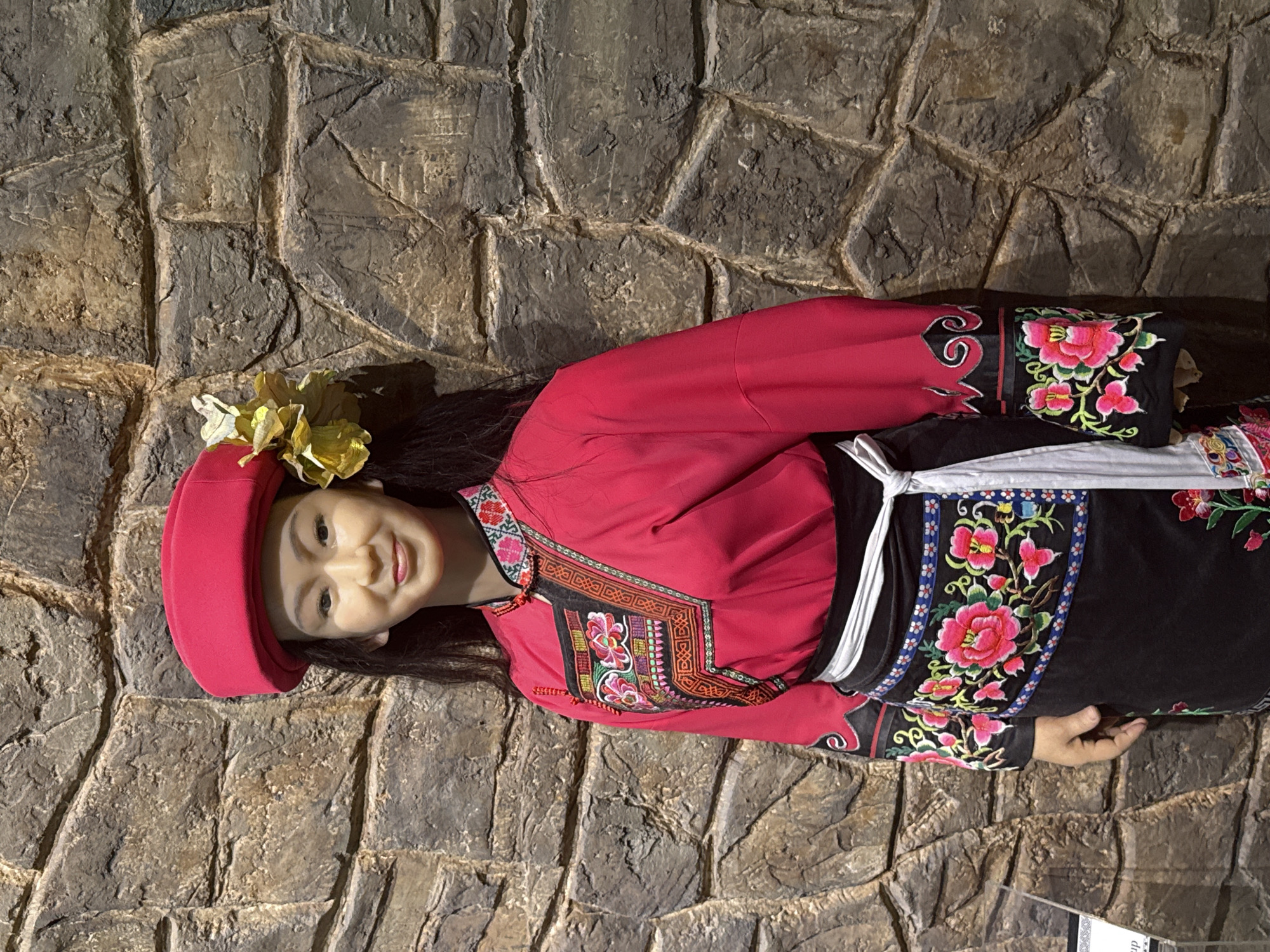 The image shows a wax figure of a young woman wearing a traditional Chinese outfit with a red top and black embroidered skirt. She is lying against a stone wall. The figure is likely an exhibit in a museum or cultural center, potentially located in the Yuzhong District of China.