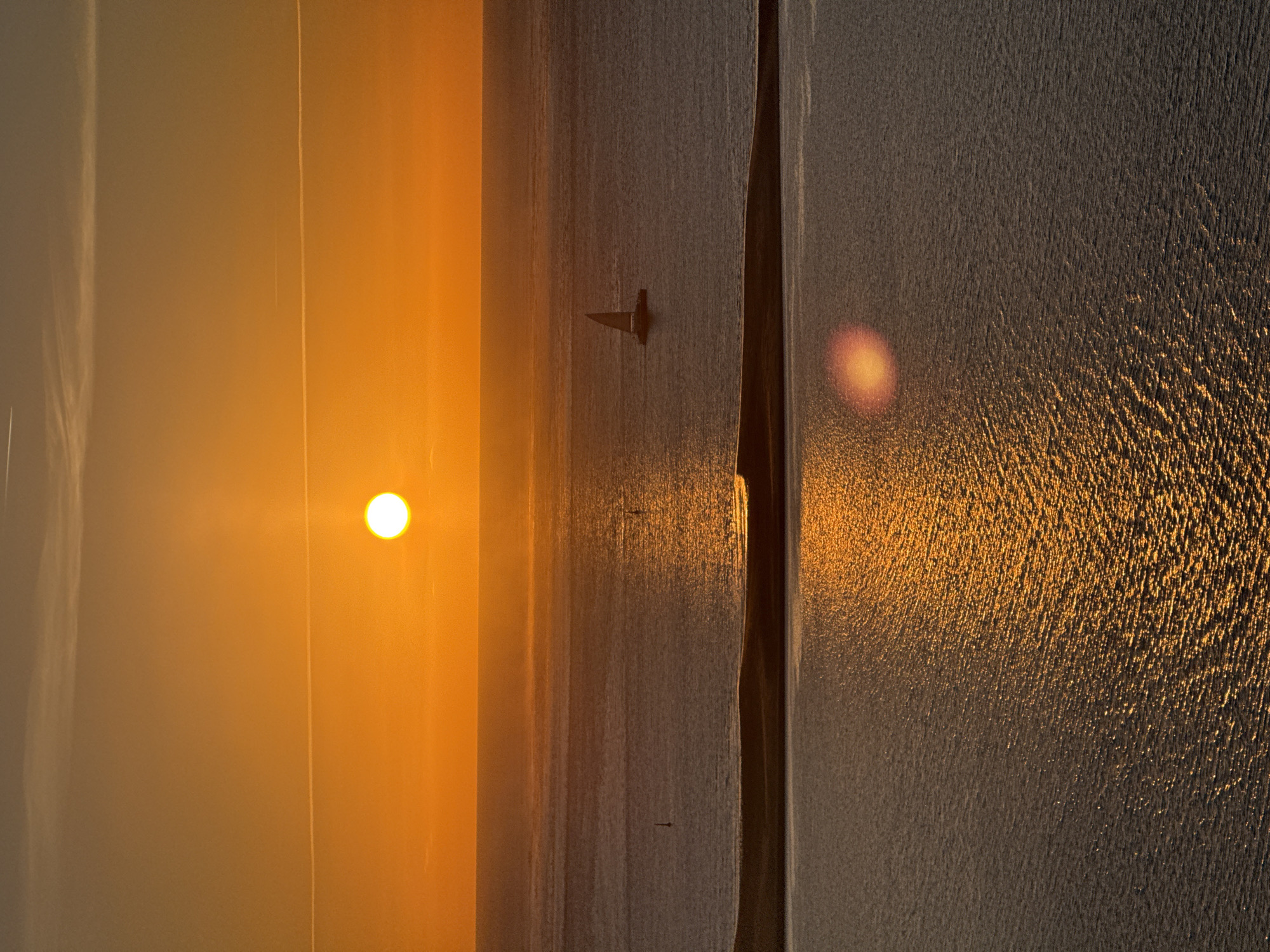 A wide-angle vertical shot captures a dramatic sunset over the ocean, likely near La Teste-de-Buch, France. The sun, a brilliant orange orb, hangs low in the sky, casting a warm glow and creating a shimmering reflection on the water's surface. The sky transitions from a deep orange near the horizon to a softer yellow and then a muted grey at the top. Two faint contrails from aircraft are visible in the sky.
In the middle ground, a narrow strip of land, possibly a sandbar or coastline, divides the ocean into two distinct bodies of water. The ocean itself is calm with gentle ripples. A lone sailboat with its sail up is visible on the further expanse of water, positioned to the right of the center. In the foreground, the water is a darker shade, reflecting the sunset colors more intensely, with a bright lens flare appearing as an orange circle on the right side. Two small buoys are faintly visible in the middle body of water. The overall atmosphere is serene and tranquil, characteristic of a coastal sunset.