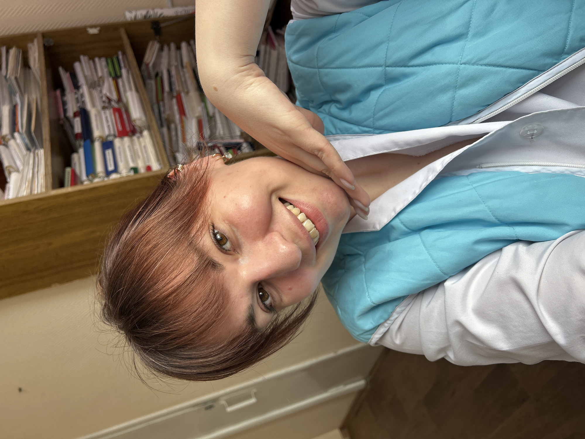 An indoor, eye-level selfie depicts a female subject, estimated to be in her 20s or 30s, situated in the city of Фоки, Russia. The woman has brown hair with reddish undertones, styled with bangs, and possesses brown eyes. She is smiling, revealing teeth. Her complexion is fair, with some redness observed on the cheeks. She wears a white collared shirt, partially unbuttoned at the neck, under a light blue, quilted vest with a zippered front. A gold-colored earring with a white drop is visible in her right ear. Her right hand is raised, with fingers gently resting against her jawline, showcasing light-colored manicured fingernails. A small mole is present on her right upper arm. The background to the subject's left reveals a light-colored wall with a partial view of a white doorframe and a wooden floor. To her right, a wooden bookshelf is visible, densely packed with numerous stacks of papers, documents, and binders across three visible shelves.