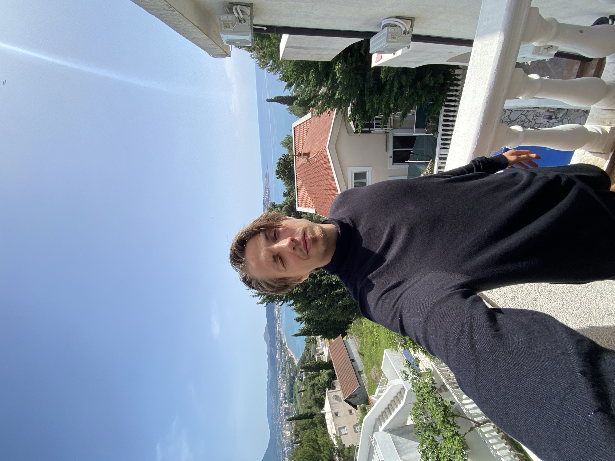 A man with blonde hair is taking a selfie on a balcony overlooking the city of Šušanj, Montenegro. He is wearing a black turtleneck sweater. In the background, there is a view of the city, the sea, and a clear blue sky. The balcony has white railings and there are trees and buildings visible in the midground. The image is rotated.