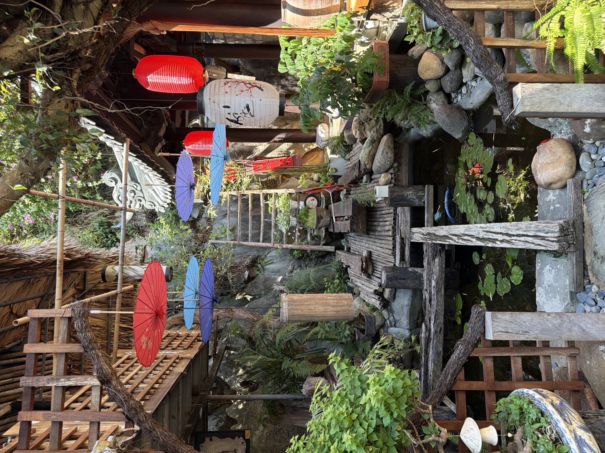 The image shows a lush, layered scene in what appears to be a garden or courtyard, likely in Đà Nẵng, Vietnam, given the cultural elements. Several tiers of wooden structures create pathways and elevated areas. Brightly colored paper lanterns and parasols are suspended overhead, adding a vibrant touch. Below, a small pond is visible, filled with aquatic plants and surrounded by rocks and greenery. Wooden steps and railings provide access to different levels. Various plants and shrubs thrive throughout the space, creating a sense of natural beauty. The composition is dense with details, showcasing a carefully designed and maintained environment.