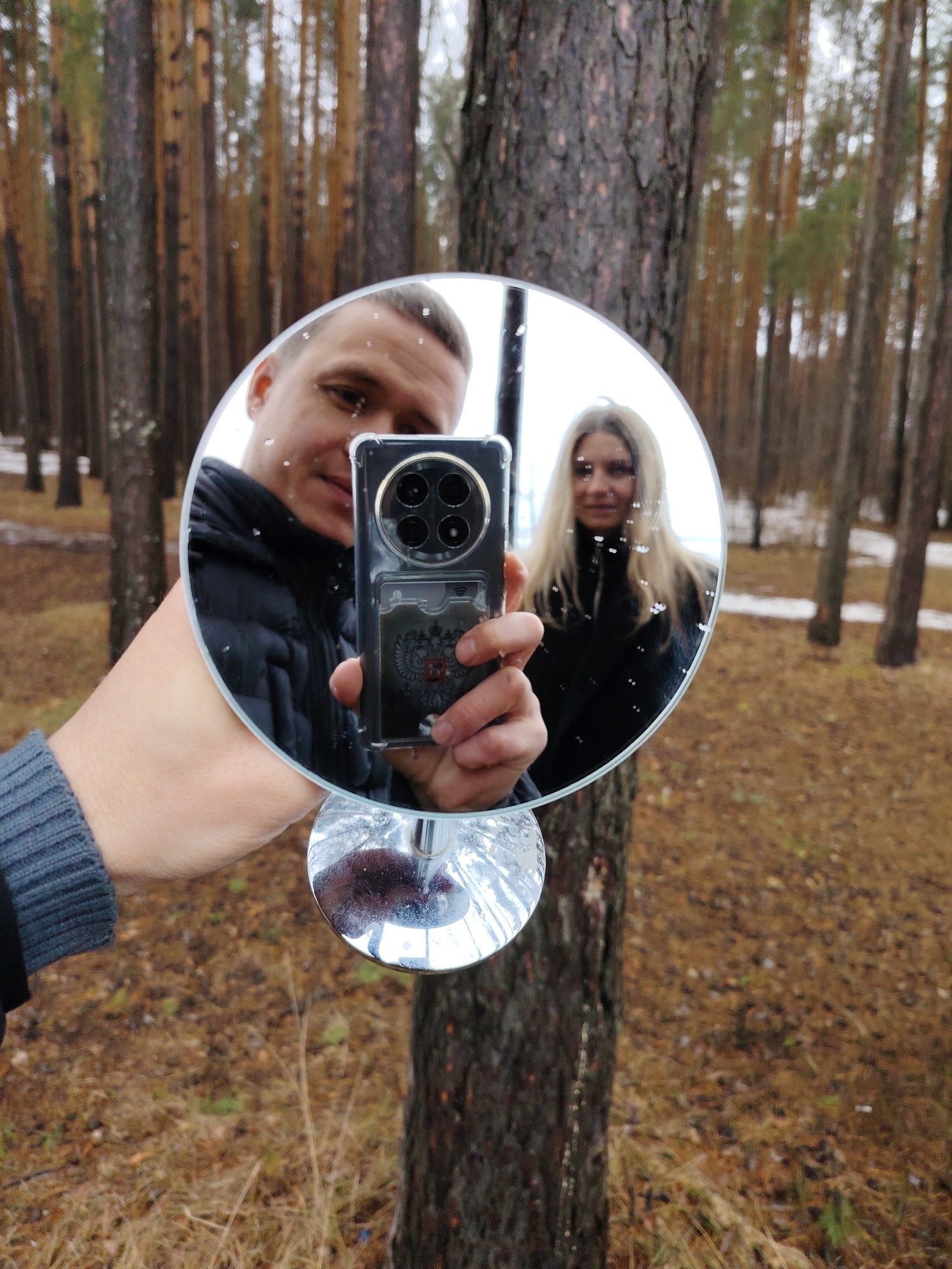 A man is holding a round mirror on a stem in front of him, reflecting his face and a woman with long blonde hair standing behind him. The man is holding a phone with a decorative case. The background shows a forest with tall trees and ground covered in dry leaves. It is located in Chaikovsky, Russia.
