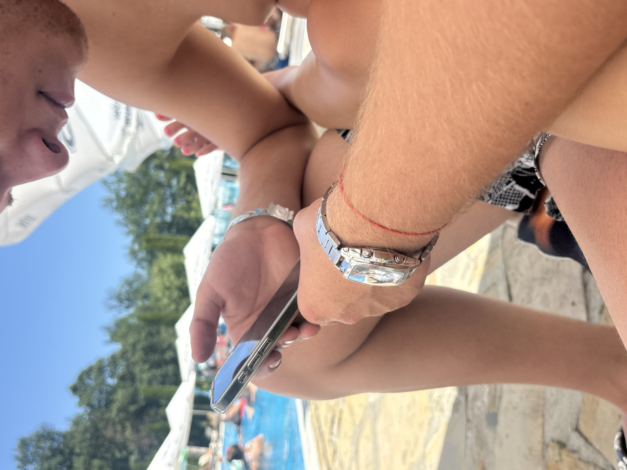 A close-up, medium shot captures a person holding a smartphone in their left hand, with their right wrist adorned with a silver watch featuring a rectangular face and a metal band. A thin red string is also tied around the same wrist. The background reveals a swimming pool with people in the water and white canopies. The scene is set outdoors on a sunny day. The individuals' lower bodies and a portion of their torsos are visible. The location is identified as Beli Osam, Bulgaria.