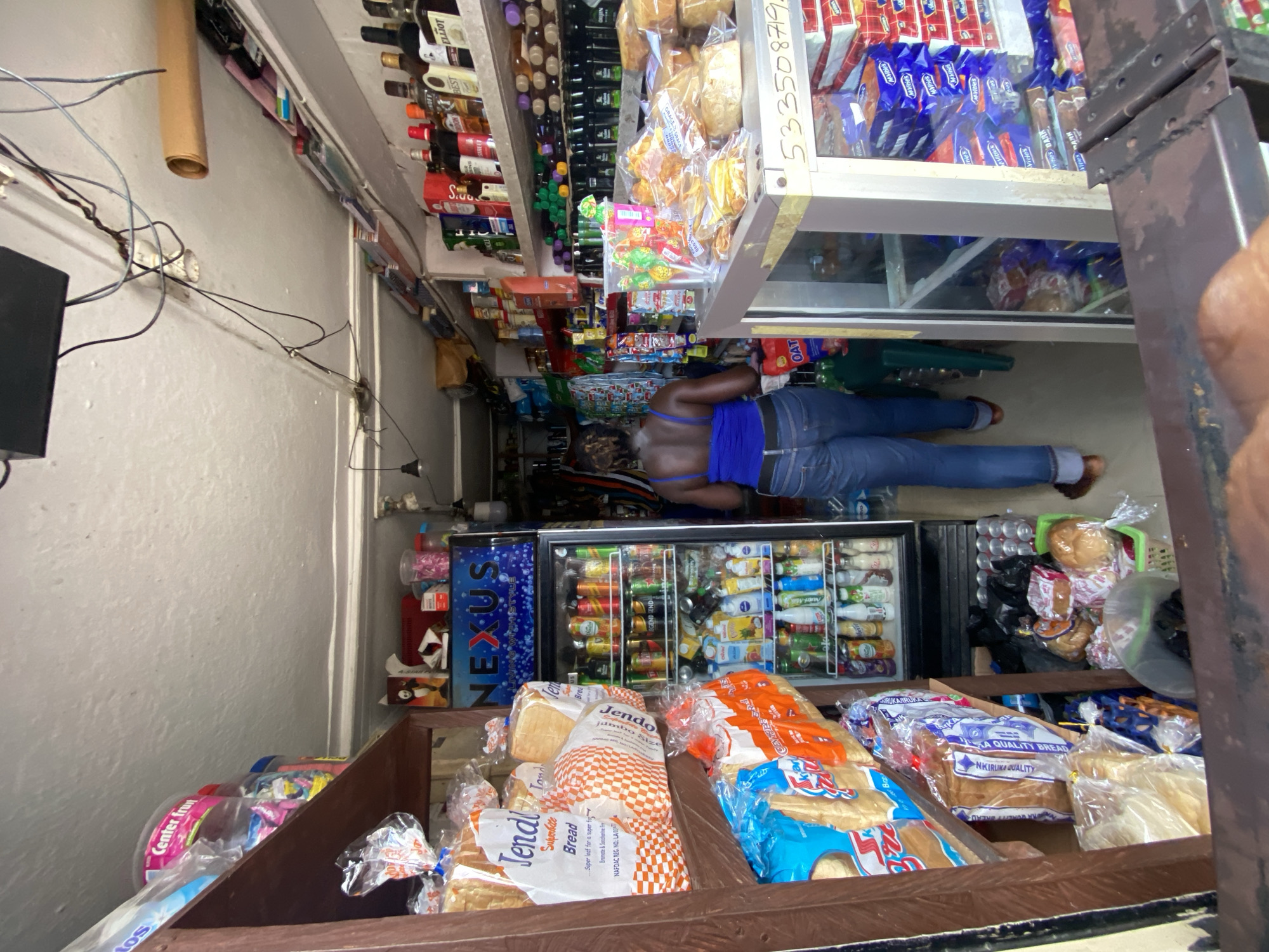 The photograph shows the interior of a small provision shop in what appears to be Ado, Nigeria. The image is taken from a low angle, looking towards the back of the store. A person, presumably a customer, is seen bending over near a glass-door refrigerator stocked with soft drinks and beverages. The shelves of the shop are packed with goods, including bottles, packaged snacks, and loaves of bread. The scene depicts a typical neighborhood store with a casual shopping environment. Visible details include the variety of products on display and the overall organization of the shop.