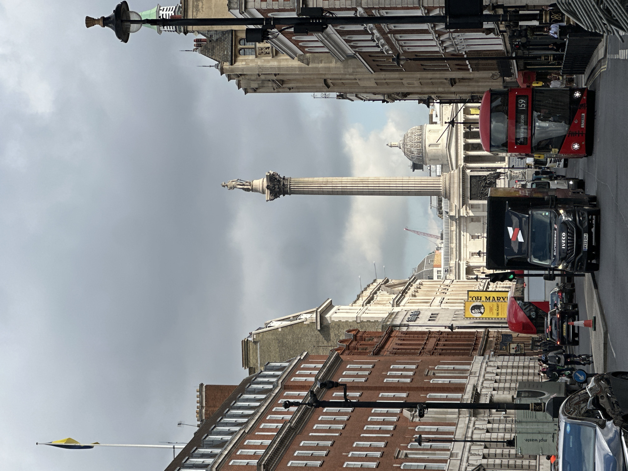 This image depicts a street scene in London, United Kingdom. Nelson's Column stands prominently, suggesting the location is Trafalgar Square. The buildings on either side of the street feature a mix of architectural styles, with brick facades and numerous windows. A red double-decker bus, specifically Route 159 to Streatham Station, and other vehicles, including a large black truck, fill the street. A yellow advertising sign with the text "OH MARY!" is visible. People are present but not clearly distinguishable. The sky is overcast with a grey sky. There are street lamps, traffic signals, and other street furniture.