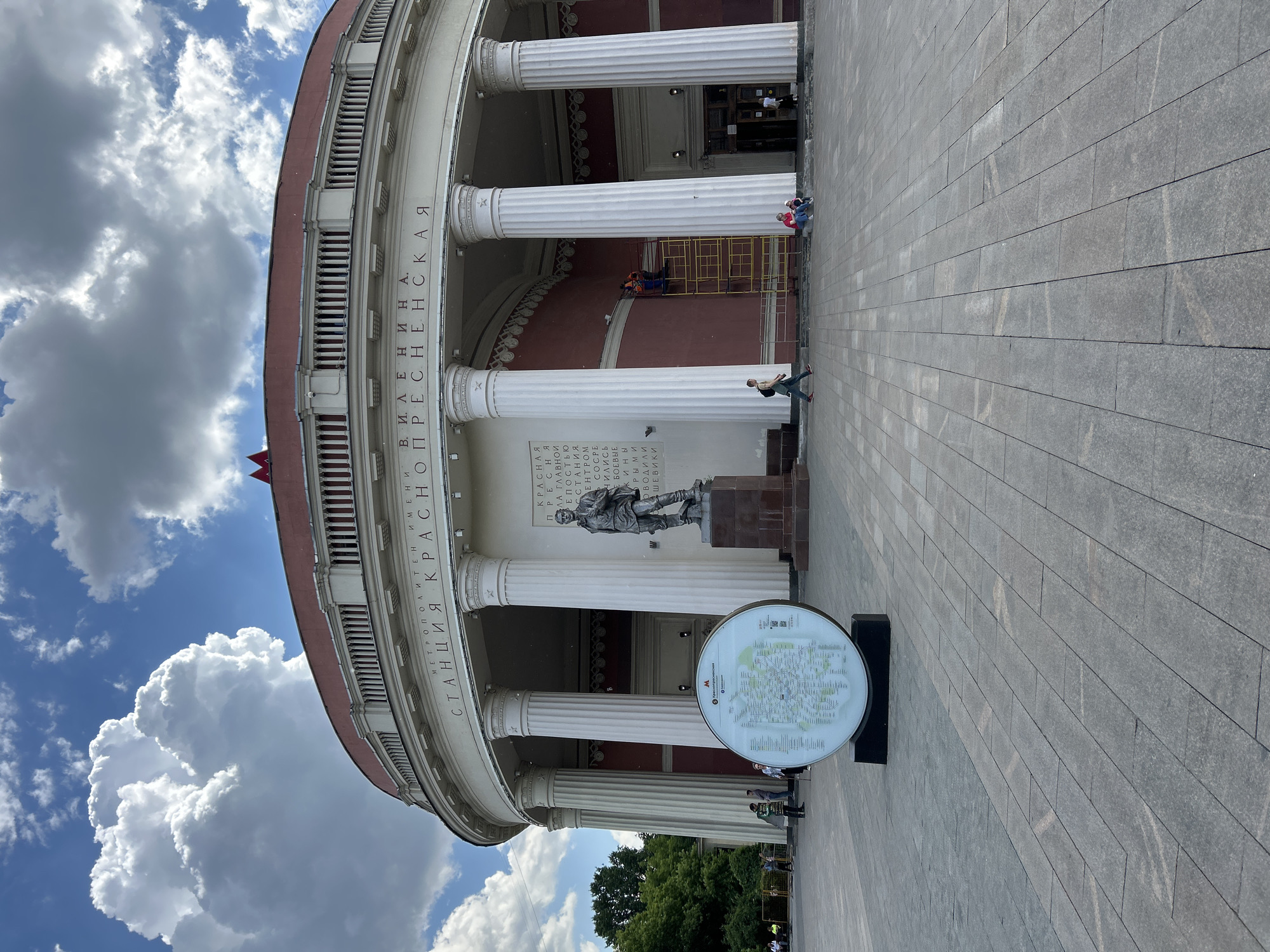 The image captures the grand, circular entrance pavilion of the Krasnopresnenskaya metro station in Moscow, Russia, under a bright, partly cloudy sky. The building exhibits a classical architectural style with a series of tall, white Doric columns supporting a red-edged entablature and an ornate balustrade.
In front of the station is a wide, light grey paved plaza. Several people are visible: one person walks across the plaza, another stands near a column, and two individuals are seated on a ledge in the shade of the portico. A worker is seen on yellow scaffolding against the red interior wall of the building, possibly performing maintenance. A dark statue of a historical figure stands on a pedestal in front of the station's main entrance, positioned beneath an inscribed plaque.
To the right, a circular, illuminated information stand displays a map, likely of the Moscow Metro system, with the station's name "M • Краснопресненская" (Krasnopresnenskaya) clearly visible.
Prominent Cyrillic text runs vertically along the façade, reading "МЕТРОПОЛИТЕН ИМЕНИ В. И. ЛЕНИНА. СТАНЦИЯ КРАСНОПРЕСНЕНСКАЯ," which translates to "Metropolitan named after V. I. Lenin. Krasnopresnenskaya Station." The plaque below the statue bears text relating to the "Krasnaya Presnya" district being the "main fortress of the uprising" where "fighting forces led by the Bolsheviks" were concentrated, referring to the 1905 Moscow uprising. The weather is sunny with large, cumulus clouds in a blue sky, indicating daytime.