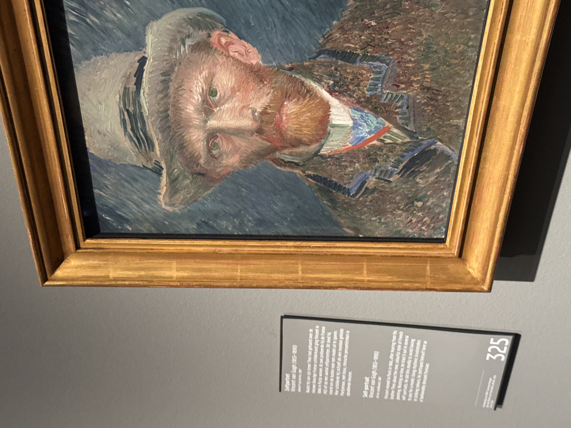 A photograph shows a framed portrait of Vincent van Gogh hanging in a museum, likely the Van Gogh Museum in Amsterdam, Netherlands. The painting depicts a self-portrait of Van Gogh wearing a hat and a jacket, rendered in his characteristic impasto style with visible brushstrokes and a palette of blues, browns, and reds. Below the painting is a small informational card labeled "325", which provides a brief description of the artwork, its context, and the artist. The wall behind the painting is painted in a neutral gray color.