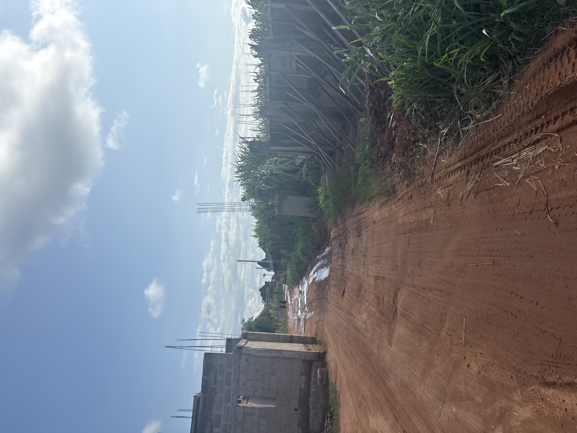 The image depicts a dirt road under a blue sky with scattered clouds. On the left side of the road, an unfinished building constructed with concrete blocks is visible, with rebar protruding from the top. Further down the road, two individuals are walking away from the camera. To the right of the road, a wooden structure, possibly a formwork for a wall, is propped up with numerous diagonal wooden beams. Tall green grass and vegetation grow on both sides of the road, particularly dense on the right. The road itself is reddish-brown and shows tire tracks from vehicles, with several puddles of water indicating recent rainfall. The scene appears to be a developing area in Orifite, Nigeria.