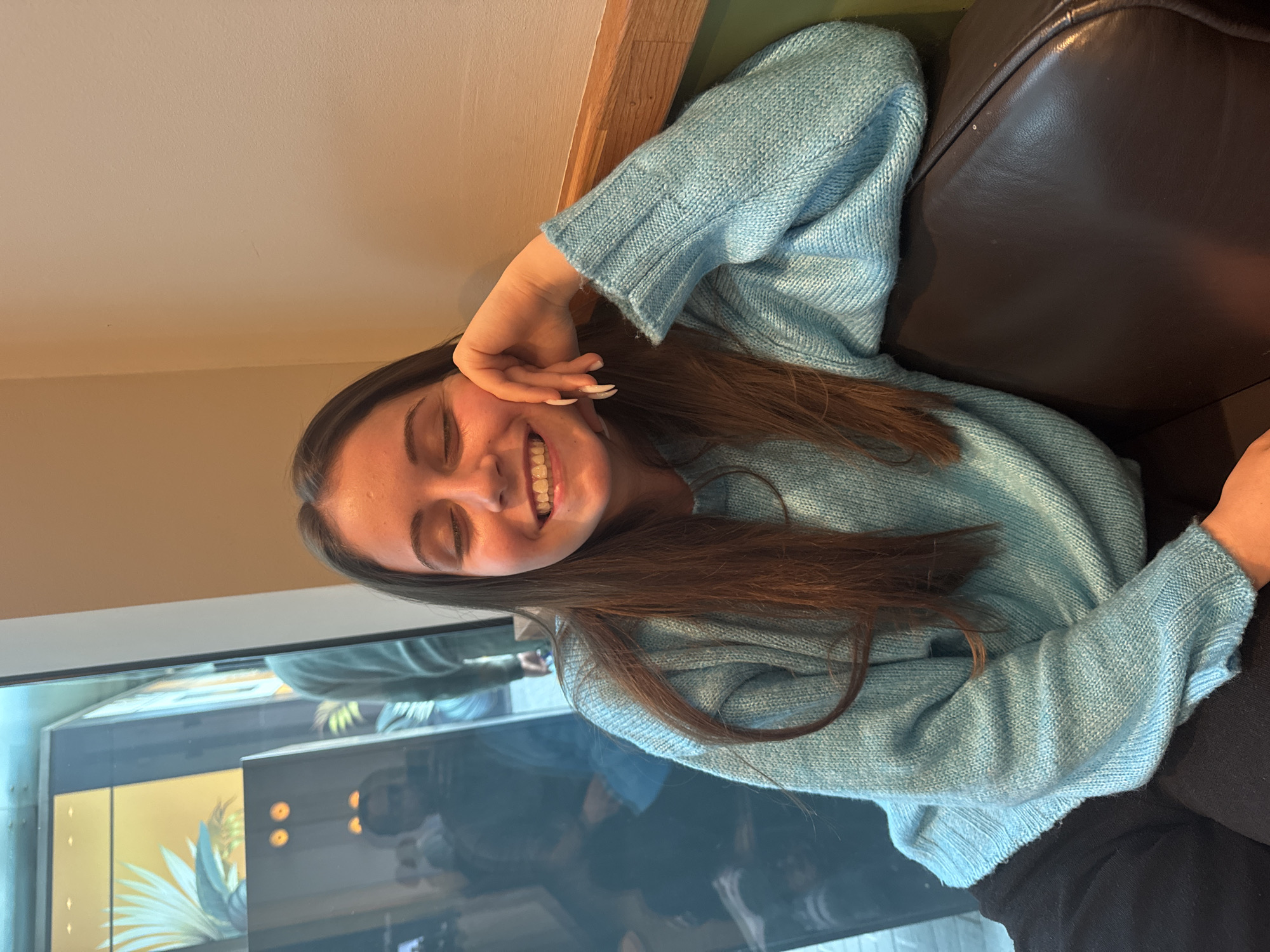 A young woman is seated on a dark brown leather couch indoors. She is wearing a light blue sweater. Her long brown hair is draped across her shoulder and chest. She is smiling with her eyes closed and her hand near her face. The location appears to be indoors, near a glass window or partition with reflections. Without any further information, determining the exact location in Burgas, Bulgaria is impossible.