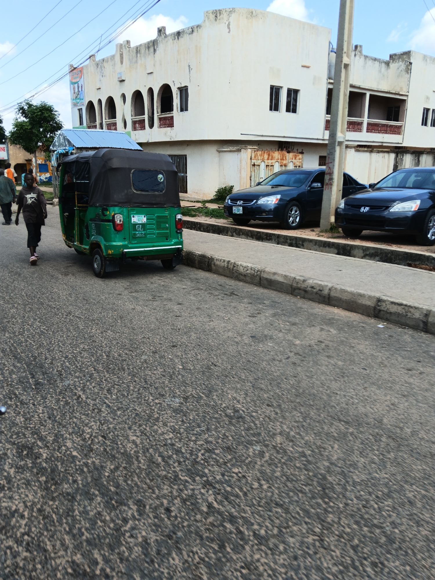 This is a photograph taken from a low angle on a street in Gombe, Nigeria. The foreground shows a textured asphalt road with a concrete curb. In the midground, a green auto-rickshaw, also known as a "Keke Napep," is parked on the right side of the road. To the left of the auto-rickshaw, a person wearing a dark t-shirt with a graphic and dark pants walks away from the camera. Further to the left, a man in a dark shirt and pants is also walking.
Behind the auto-rickshaw, two dark-colored sedans are parked on the side of the road. The car closer to the foreground has a license plate that reads "KC-33B1A".
In the background, a two-story building with a pale yellow facade dominates the scene. The building has arched windows and balconies with decorative railings. A large banner with portraits and text is visible on the left side of the building. The sky above is blue with scattered white clouds. A utility pole is visible to the right of the sedans. The scene appears to be a typical street view in an urban area of Gombe.