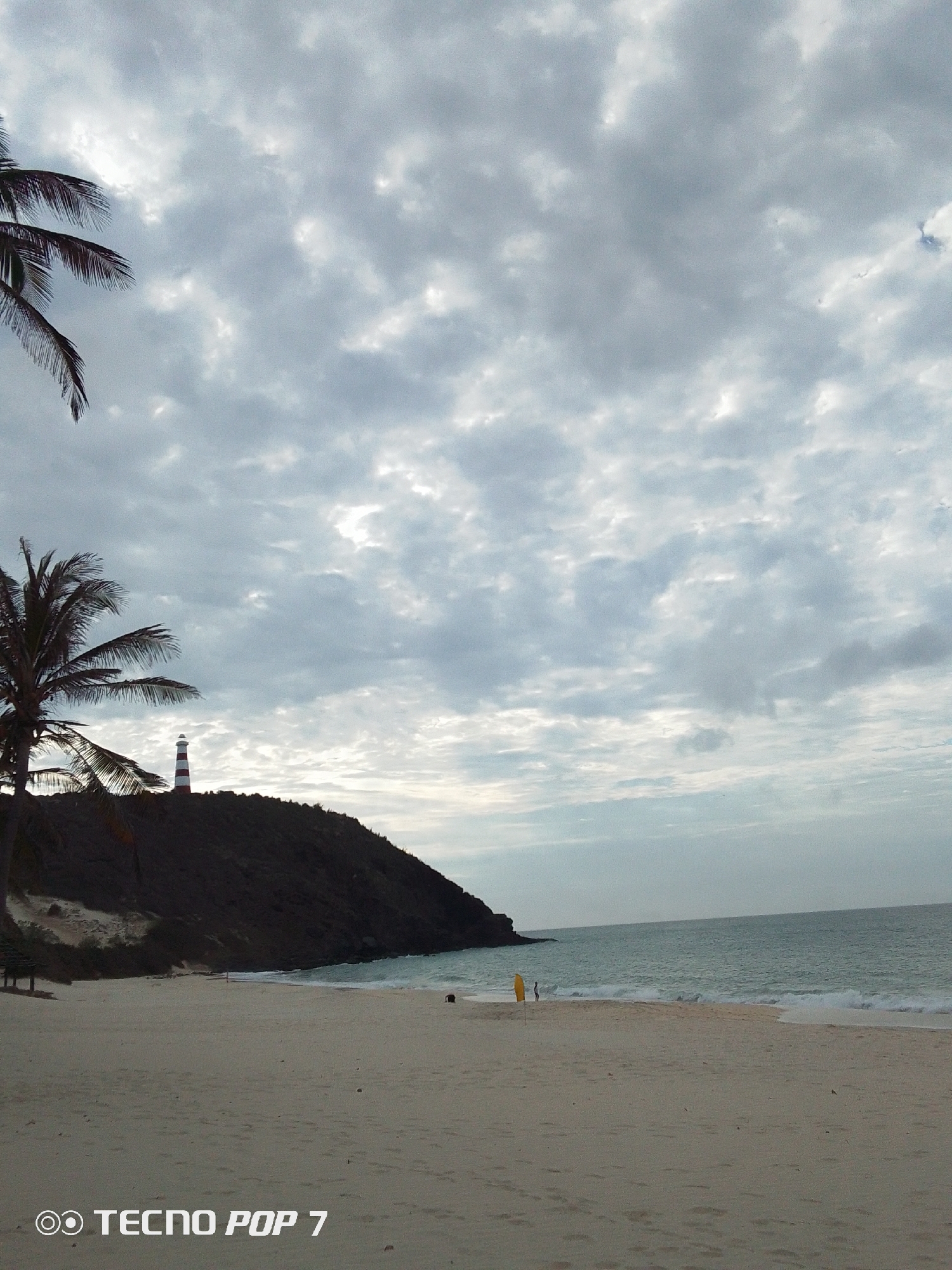 This photograph depicts a beach scene near Pedro González, Venezuela, under a cloudy sky. A beach stretches across the foreground, revealing patterns in the sand. The sea meets the sand with small waves. On the beach, there's a yellow object and a person standing. In the midground, a hill with a lighthouse stands next to some palm trees. The sky is overcast. The watermark "TECNNO POP 7" is visible in the bottom left corner.