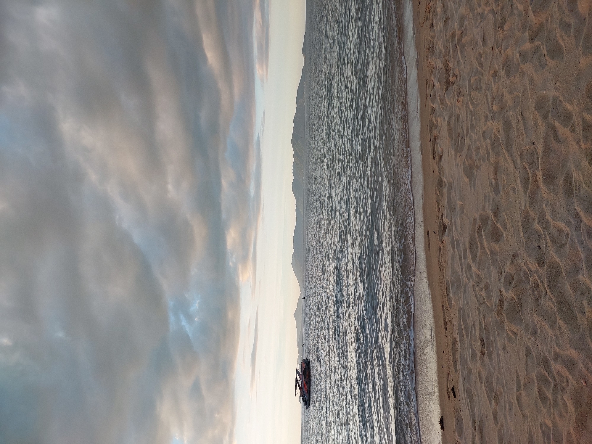 This is a vertical, eye-level shot of a serene beach scene at Baan Mae Nam, Thailand, likely during dusk. The sky is filled with soft, diffused clouds that are painted with hues of pale yellow, pink, and gray, hinting at the fading sunlight. Below the sky, a distant mountain range is silhouetted against the pale horizon.
The middle ground is dominated by the calm, rippling surface of the ocean. The water is a dark, muted blue-gray, reflecting the soft light of the sky. Along the water's edge, a thin line of white foam marks the gentle lapping of waves on the shore.
In the water, a single object is visible: a jet ski. It appears to be a dark color, possibly with red accents, and it is stationary. A person, or possibly two, can be seen on the jet ski, but they are small and indistinct from this distance.
The foreground is occupied by a sandy beach. The sand is a warm, light brown and is textured with numerous small waves and ripples, likely created by the wind. There are also a few scattered footprints visible in the sand, indicating recent human presence. The overall mood of the image is peaceful and tranquil, with the soft lighting and gentle elements creating a sense of calm. No text is visible in the image.