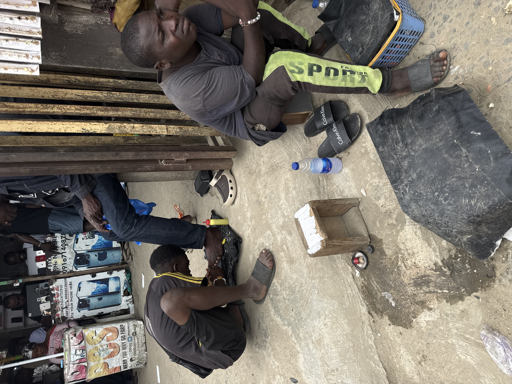 In Lekki, Nigeria, a man is crouched on a concrete ground, appearing to be working on another person's feet, possibly a pedicure or foot treatment. He wears a black t-shirt and shorts, with a yellow stripe on his shorts. The person whose feet are being worked on is wearing a dark t-shirt and dark pants with a neon green stripe. They are seated on a low wooden stool. Scattered around them on the ground are several pairs of sandals, including a pair of black Calvin Klein slides, a yellow bottle, and a blue plastic basket containing various items. In the background, a kiosk with advertisements for phone repairs and sales is visible, with several individuals standing and seated behind it. The scene suggests a bustling outdoor commercial area.