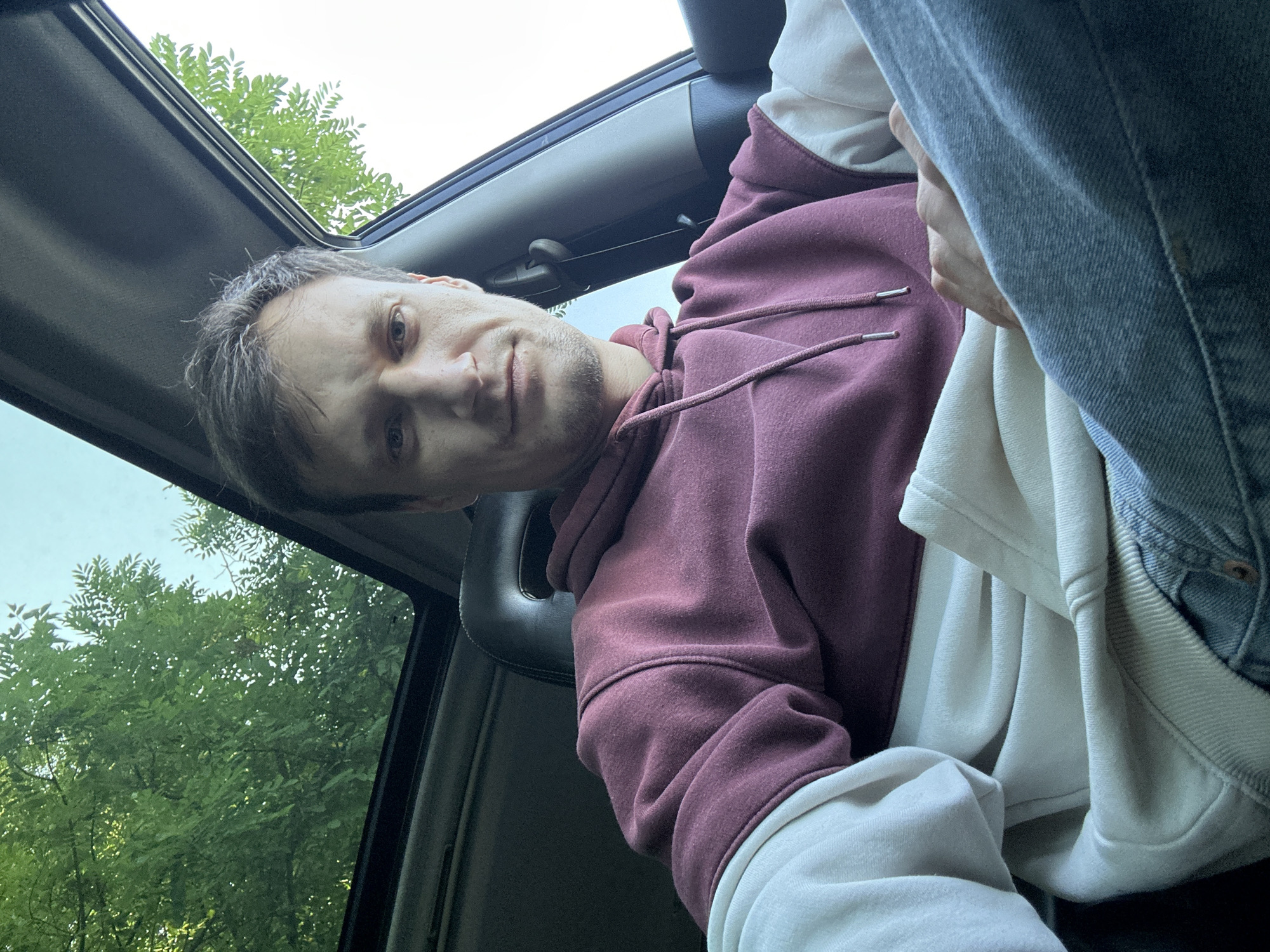 A man is seated inside a car with a sunroof open, looking directly at the camera. He is wearing a two-tone hooded sweatshirt, with the upper portion being maroon and the lower sleeves and body a cream color. He is also wearing blue jeans. Green leaves and a clear sky are visible through the sunroof and the car window, suggesting an outdoor setting possibly during the day. The image appears to be a selfie taken from the driver's seat. The location is not identifiable from the visual information provided in the image.