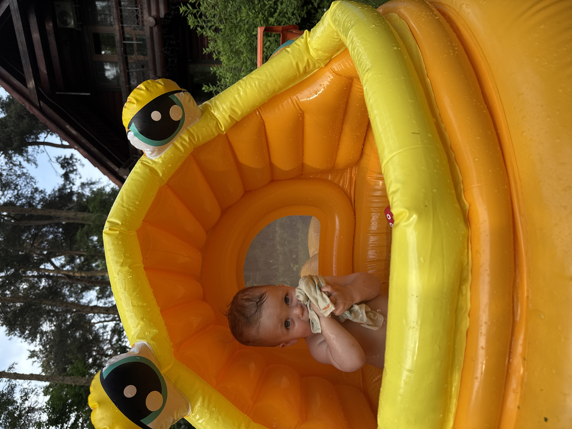 A close-up shot shows a baby inside a yellow and orange inflatable pool. The pool is located outdoors, in what appears to be a garden setting near a wooden building and trees. The baby is holding a light-colored cloth, possibly wiping its face. The picture was taken in Tomilino, Russia.