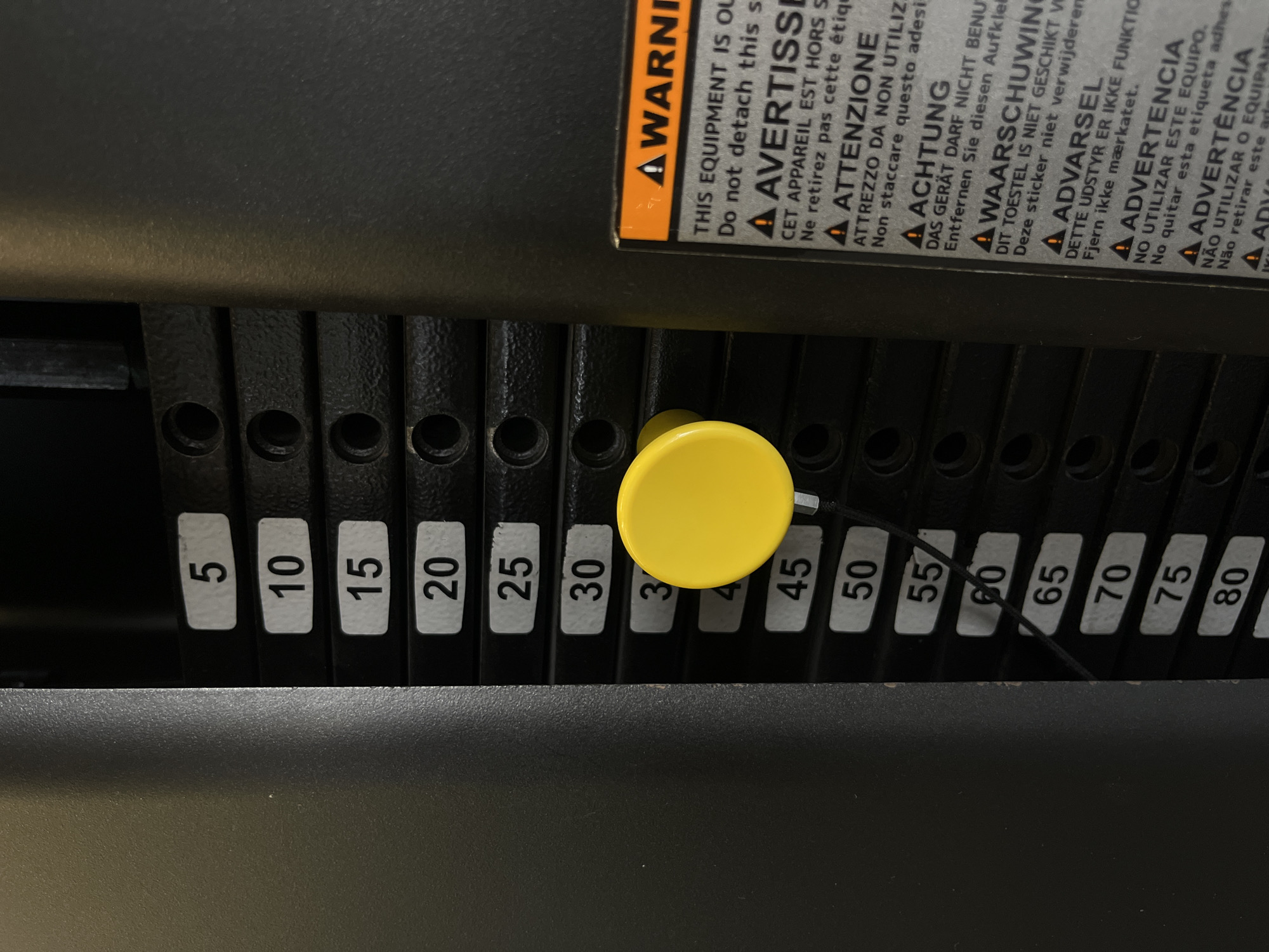 A close-up shot depicts the weight stack on a weightlifting machine. The weights are black with white numbers indicating their value in increments of 5, ranging from 5 to 80. A bright yellow selector pin is inserted into the 35-pound weight, indicating the current weight setting. In the background, there is a warning label adhered to the machine frame with multilingual text. The environment appears to be an indoor gym setting.