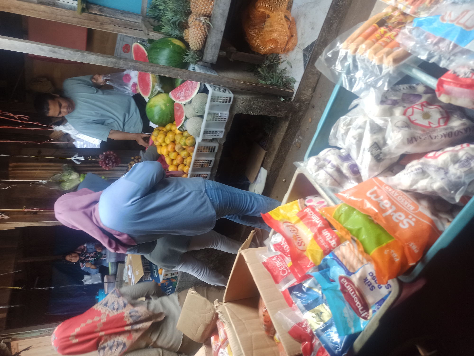 The image depicts a street market scene in Trisinar, Indonesia. In the foreground, several plastic bags containing food items, possibly processed meats or snacks, are arranged in a blue plastic container. Some of these bags are labeled with Indonesian text, such as "Indomina Stik Rasa" and "salam BAKSO AYAM". Behind this, to the left, is a cardboard box also containing packaged food items.
In the mid-ground, a woman wearing a red and patterned headscarf stands facing away from the camera, and another woman in a blue headscarf and a light blue long-sleeved shirt is also visible. Further back, a man in a light green t-shirt is holding a plastic bag and appears to be interacting with a customer. He is standing behind a stall displaying various fruits including watermelons (some cut open), oranges, and pineapples. Several white plastic baskets hold the fruits.
In the background, a woman with a headscarf and patterned top is partially visible, standing behind a counter with blue containers. The environment suggests an outdoor market with makeshift stalls constructed from wood and possibly metal sheeting. The overall impression is of a busy, local market.