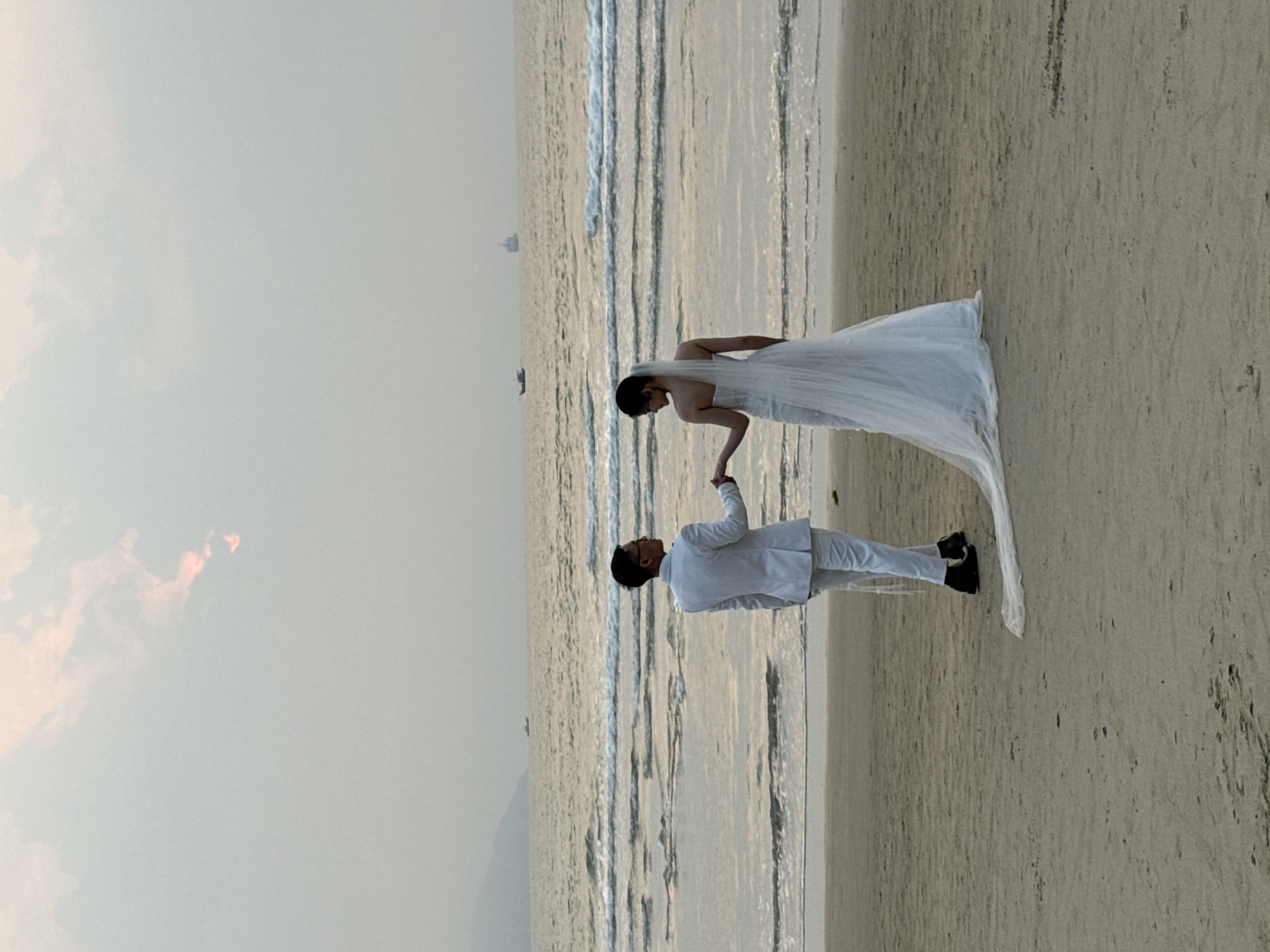A wedding couple poses on a beach. The bride wears a white wedding gown with a long train, and the groom wears a white suit with dark shoes. They are holding hands. The location is Đà Nẵng, Vietnam, specifically a beach with calm waves. The sky is overcast.