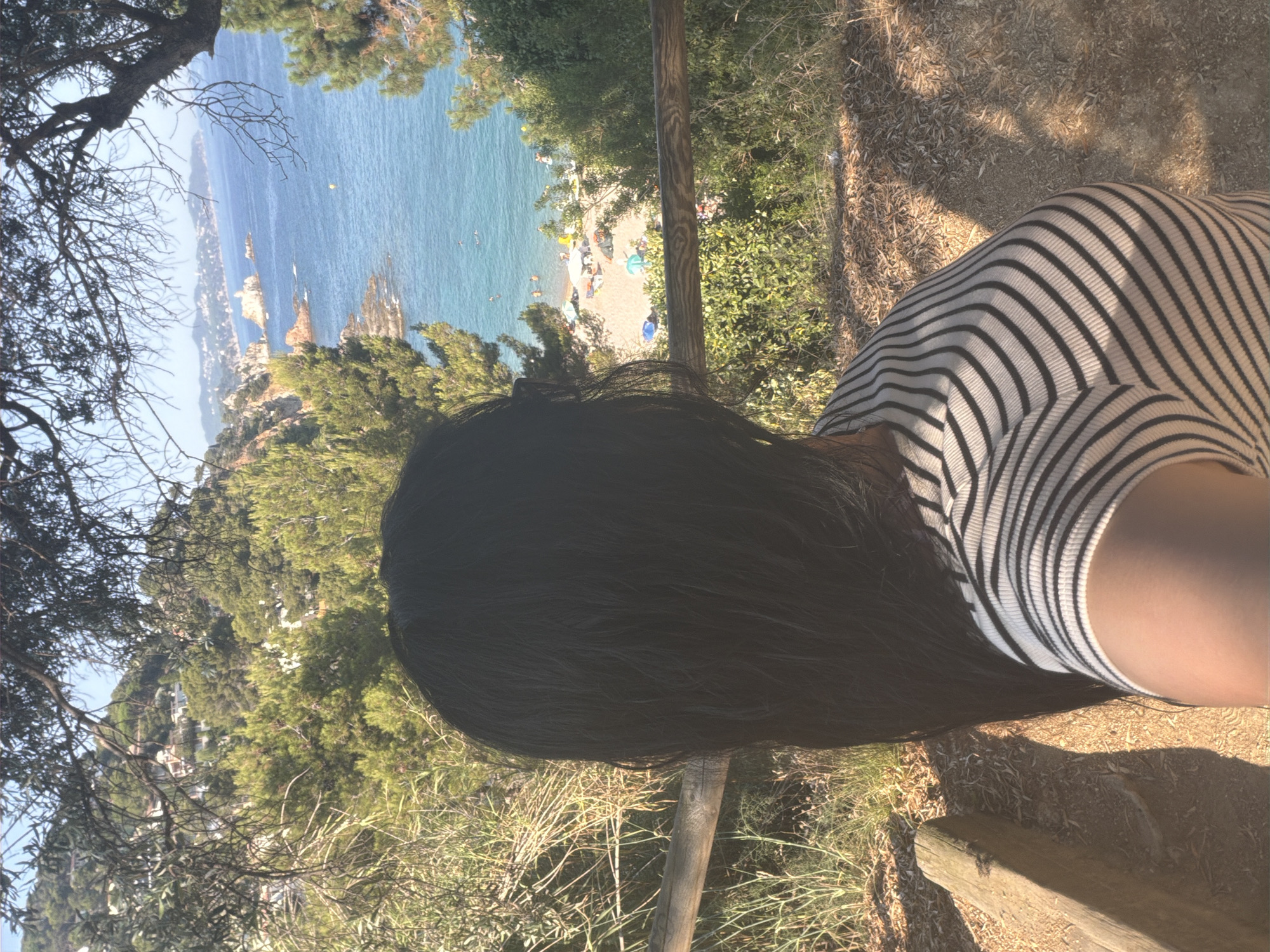 The image shows a woman from behind, facing a scenic view of the coastline. She has long, dark hair and is wearing a black and white striped shirt. She is standing on a dirt path with dry vegetation and a wooden fence in the foreground. Beyond the fence, the blue sea is visible, with a beach dotted with umbrellas and people in the distance. Rocky outcrops and lush green trees frame the view of the sea and the town nestled on the hills. The scene appears to be from a vantage point overlooking the coast of Blanes, Spain, possibly near the botanical gardens or a viewpoint along the Camí de Ronda coastal path. The sky is clear and blue, indicating a sunny day.