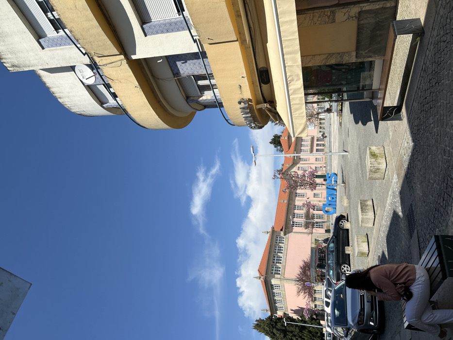 The image displays an urban square in Chaves, Portugal, under a clear blue sky with minimal white clouds. In the foreground, a female with dark hair, wearing a light pink long-sleeved top and white trousers, sits on a slatted wooden bench with dark metal supports, positioned in partial shadow on a cobblestone-like surface. Her right arm is raised towards her face.
To the left, several vehicles are parked parallel to a curb, including a silver Volkswagen Polo facing right and a black station wagon behind it. The central paved area of the square contains multiple rough-hewn grey concrete blocks and a large, blue, three-dimensional sign spelling "Chaves." Two tall, grey streetlights are also visible.
In the background, a large, multi-story building with a pink facade, numerous windows, and an orange tiled roof stands prominently, flanked by trees with pink blossoms. To the right, a curved, multi-story building with a yellow/beige facade and white sections occupies the frame. This building features balconies with blue and white patterned tile railings, white roll-down shutters on the windows, and a satellite dish mounted on an upper balcony. The ground floor displays a light-colored awning over a shop window, with a partially visible sign above reading "CAMISARIA S" and a street sign below identifying "Rua Candido Sotto Mayor."