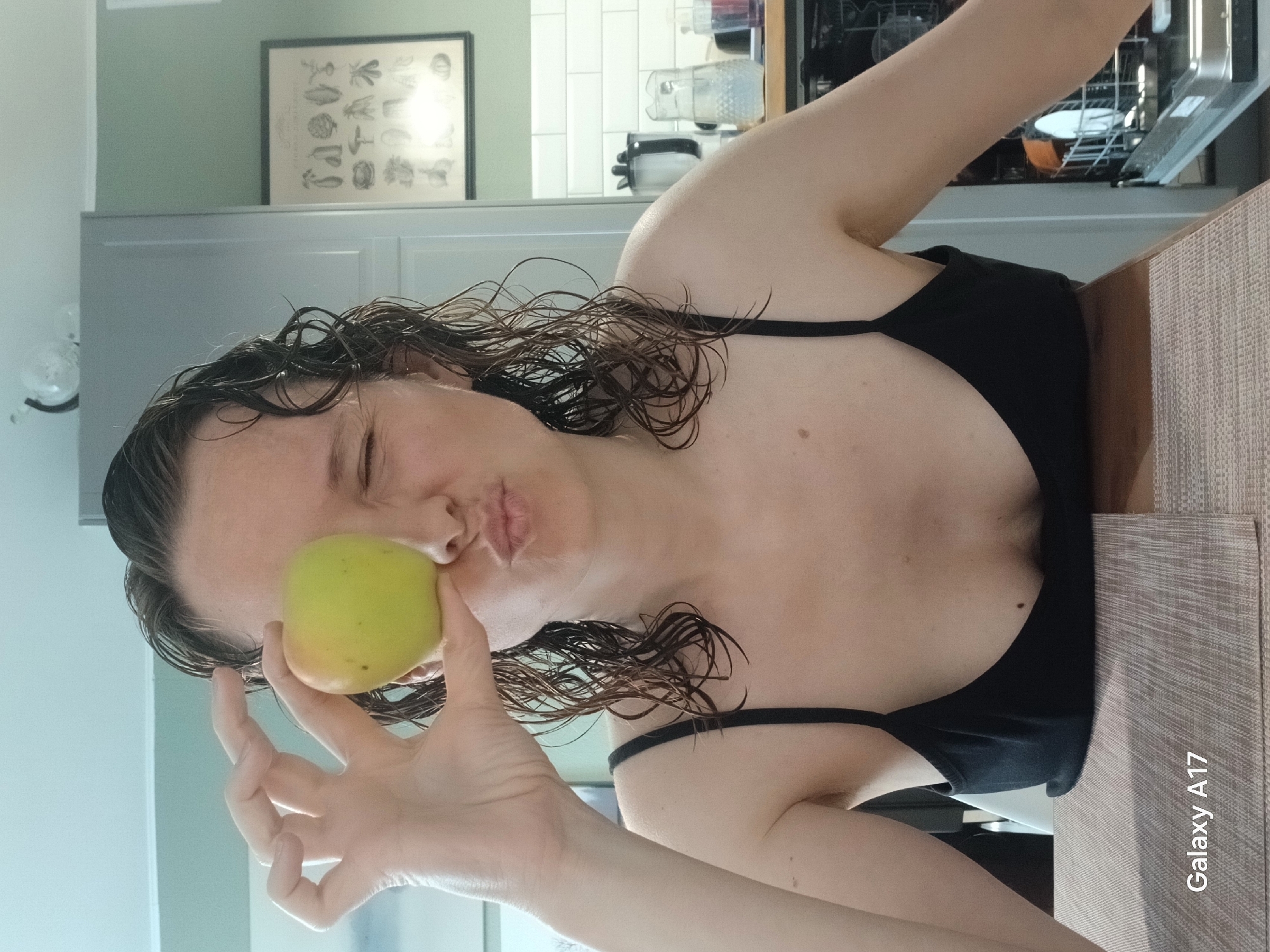 A woman is posing indoors, holding a green apple to her face as if kissing it. She has wet, curly brown hair and is wearing a black top with thin straps. The background includes a kitchen with light green walls, white cabinets, a framed botanical print, a dishwasher, and various kitchen appliances. The woman is sitting at a table with a textured surface. The bottom right corner has the text "Galaxy A17". There is no indication that this is in Yekaterinburg, Russia.