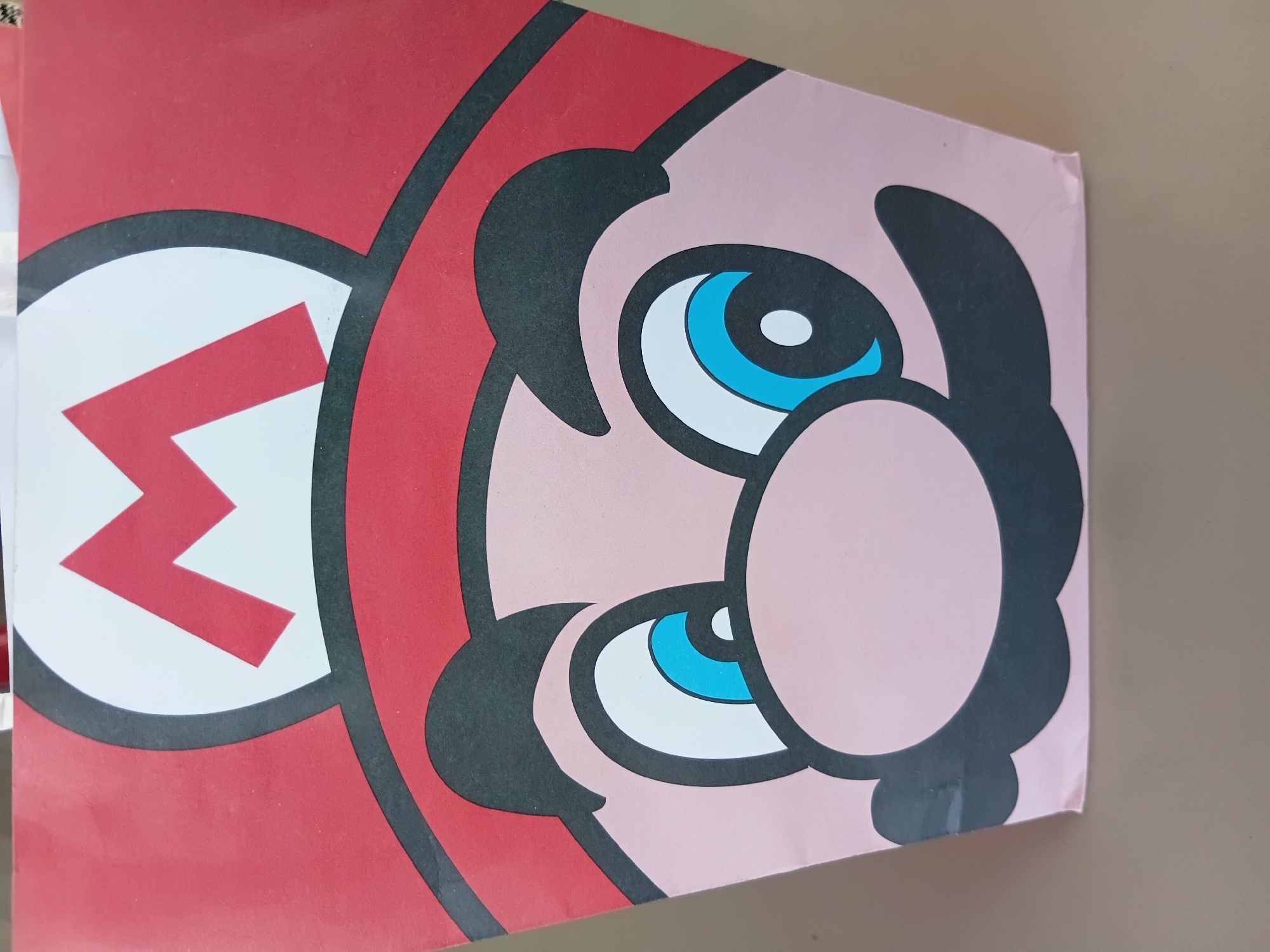 The image is a close-up of a printed representation of Mario, the iconic Nintendo character. The focus is on his face, including his hat with the "M" logo, his eyes, nose, and the black outline of his features. The colors are vibrant, typical of Mario's design, with red, white, pink, blue, and black. The photograph appears to have been taken indoors. The image does not contain any information about the location being in Lhokseumawe, Indonesia.