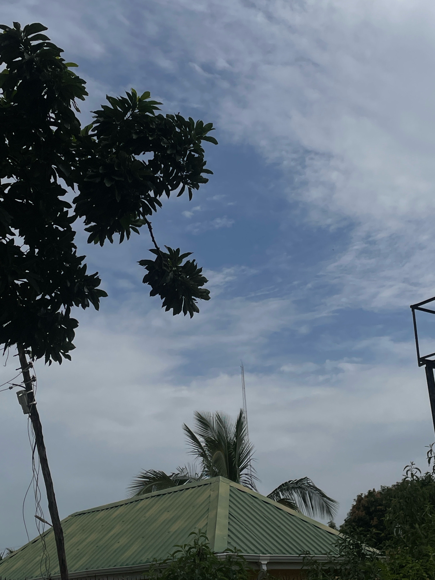 The image depicts a partly cloudy sky with a few wispy clouds scattered across a pale blue expanse. In the foreground, the dark silhouette of a tree with broad leaves occupies the left side of the frame. A wooden utility pole, with electrical wires attached, stands vertically near the tree. Below this, a house with a green, corrugated metal roof is visible. The roof has gutters along its edge. Behind the house, palm fronds and other greenery can be seen. In the distance, a tall, thin antenna or spire rises into the sky. A black metal structure, possibly part of a basketball hoop or similar frame, is visible on the far right. The scene appears to be set in an outdoor environment, likely a residential area within the city of Gura Top, Nigeria.