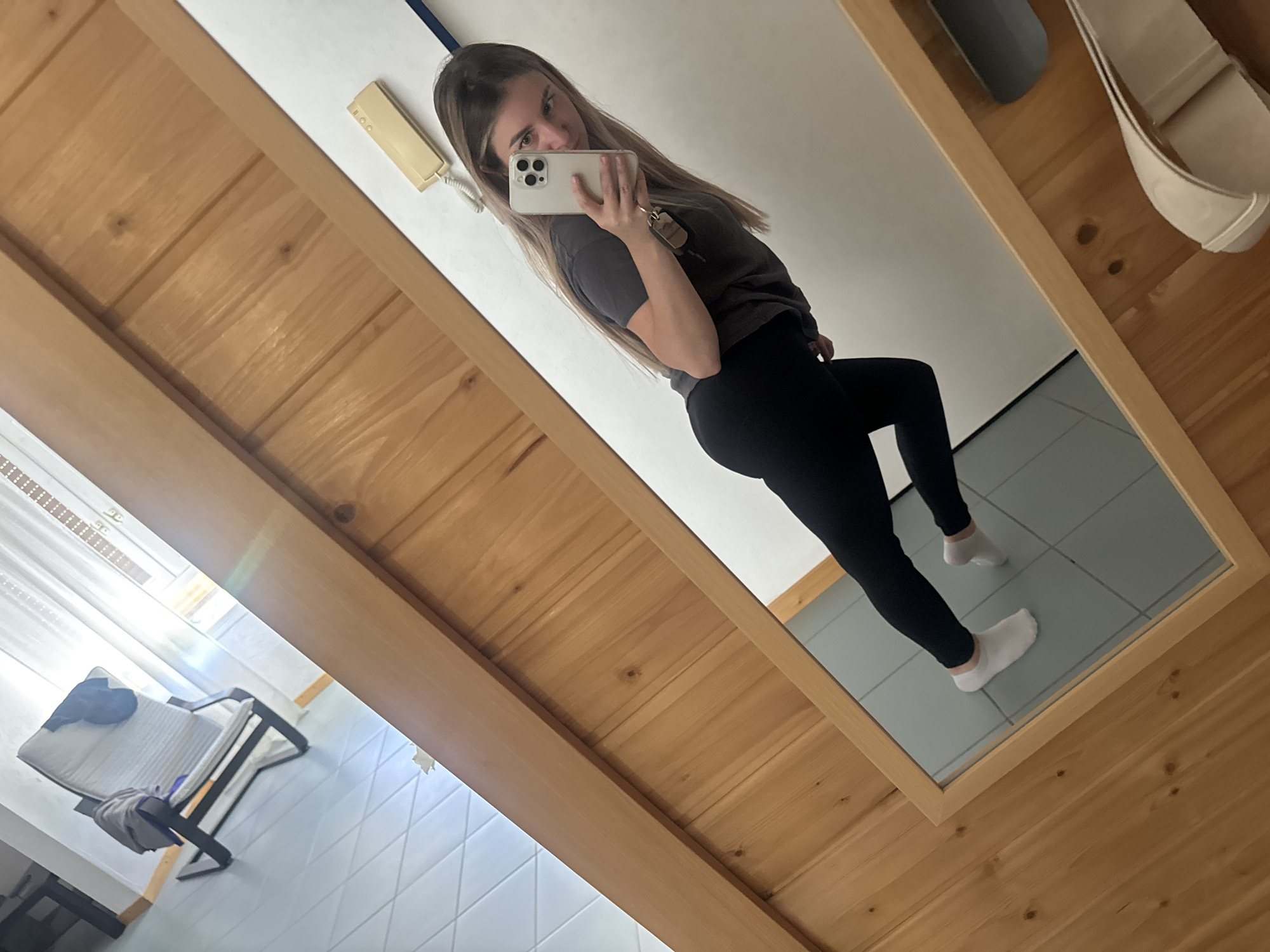 The image shows a young woman taking a mirror selfie in what appears to be a room with wooden paneled walls and a tiled floor. She is wearing a dark t-shirt, black leggings, and white socks. She is holding a smartphone in her right hand and looking at her reflection. In the background, a chair with a blanket on it is visible, along with a window with blinds. A landline phone is mounted on the wall. The scene is indoors, and there is no indication of the city of Saint-Claude, France.